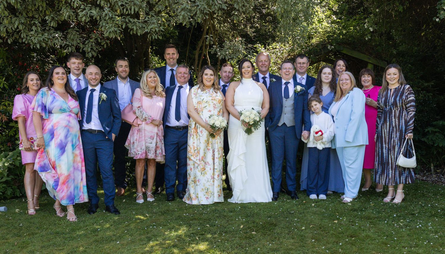 Large wedding party poses for group photo outdoors in lush garden setting.