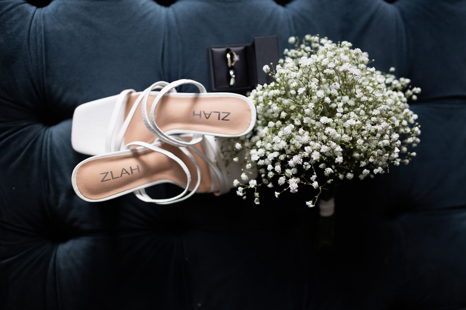 White bridal shoes and a baby's breath bouquet arranged on a dark tufted couch.