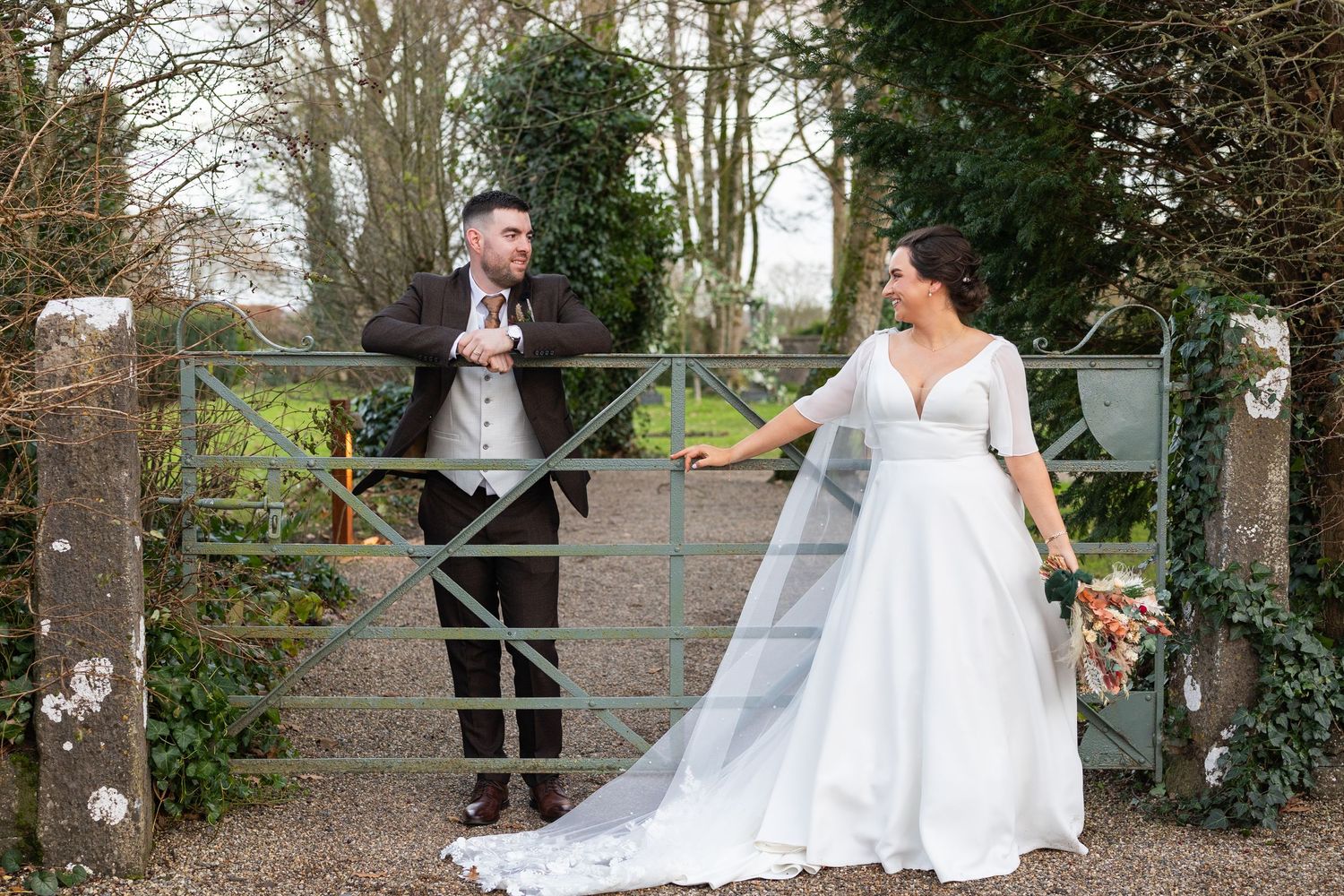 Bride and groom posing on opposite sides of a rustic gate, bride wearing a flowing veil.