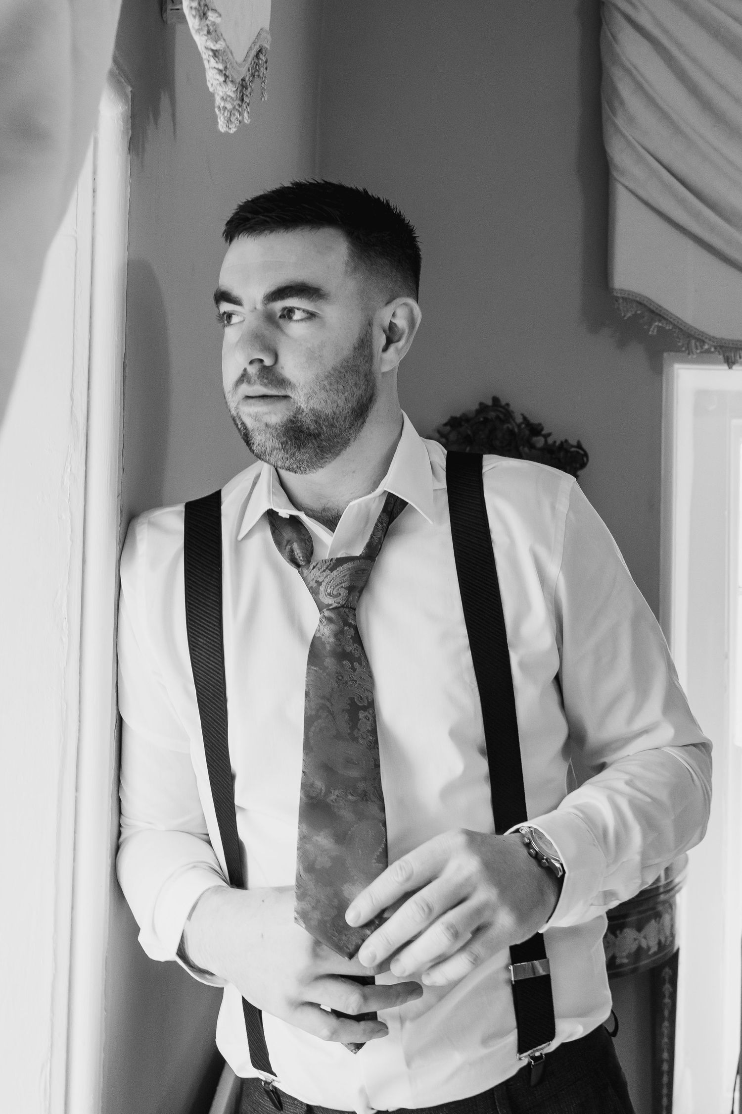 Black and white portrait of a groom in white shirt, suspenders and tie preparing for his wedding day.