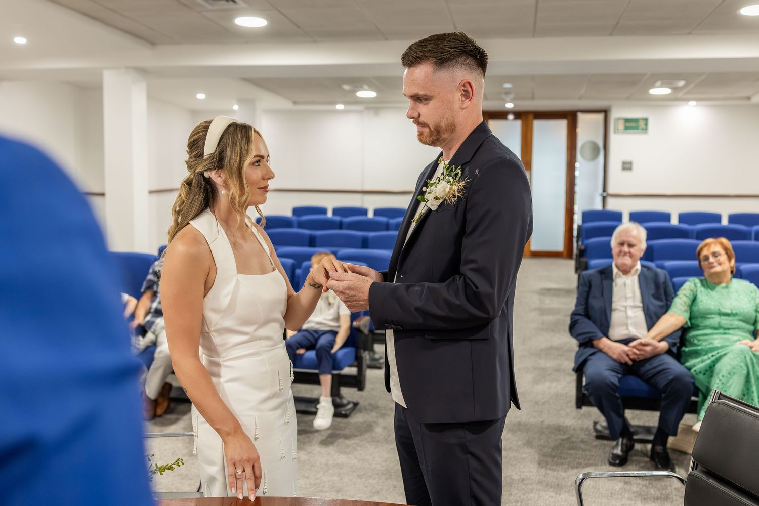 Couple exchanging rings during a civil wedding ceremony in a registry office with guests seated in blue chairs.