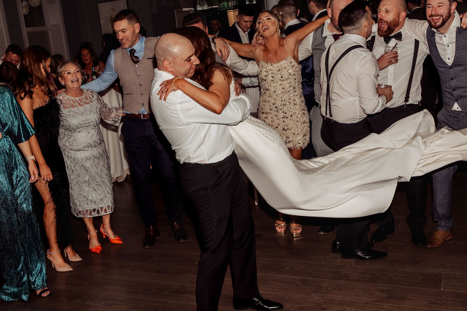 Groom lifts bride in wedding dress on dance floor surrounded by celebrating guests at reception.