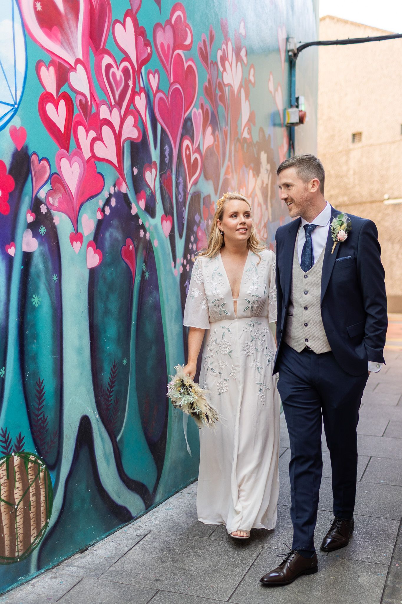 Wedding couple posing against vibrant street art mural with pink hearts and teal background.