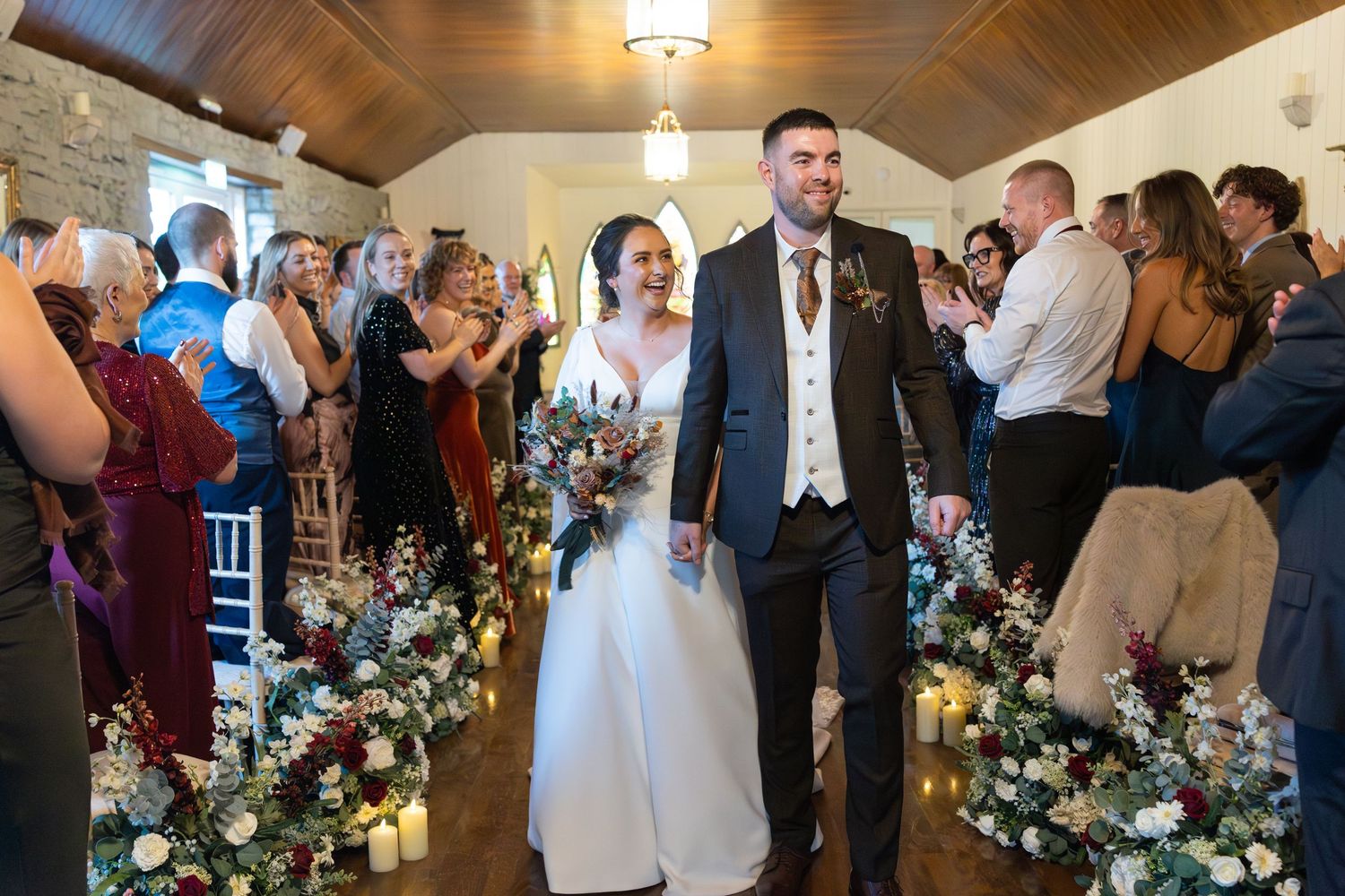 Bride and groom walk down the aisle together, surrounded by floral arrangements and cheering wedding guests.