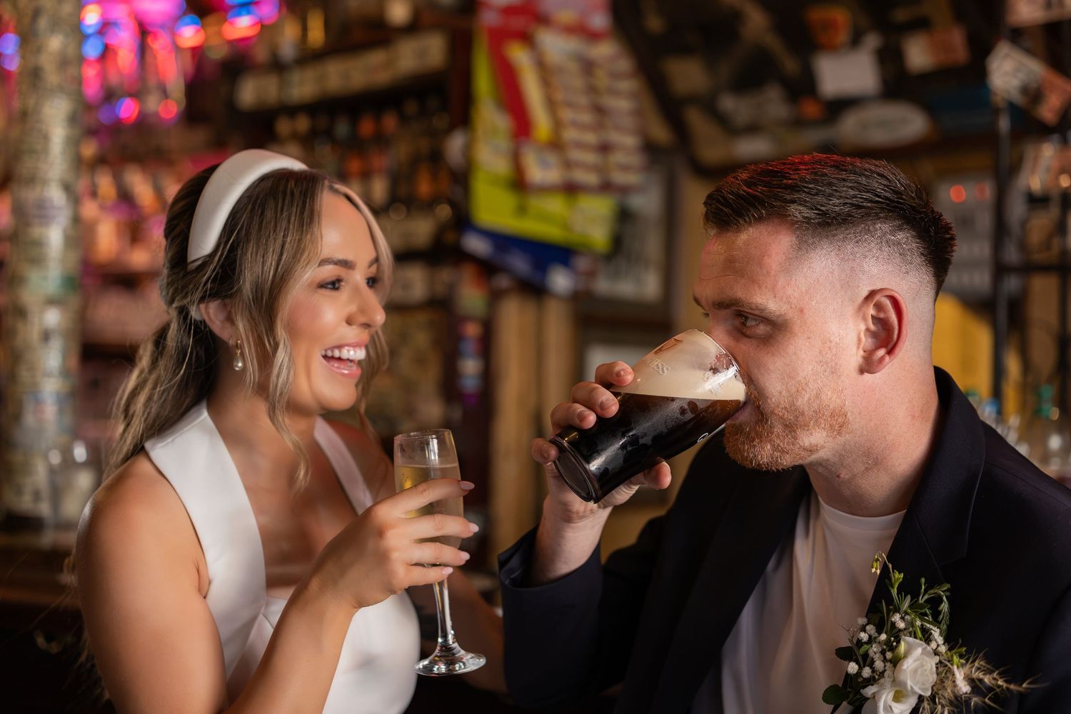 Bride and groom celebrate at a bar, she holds champagne while he drinks a dark beer, both smiling joyfully.