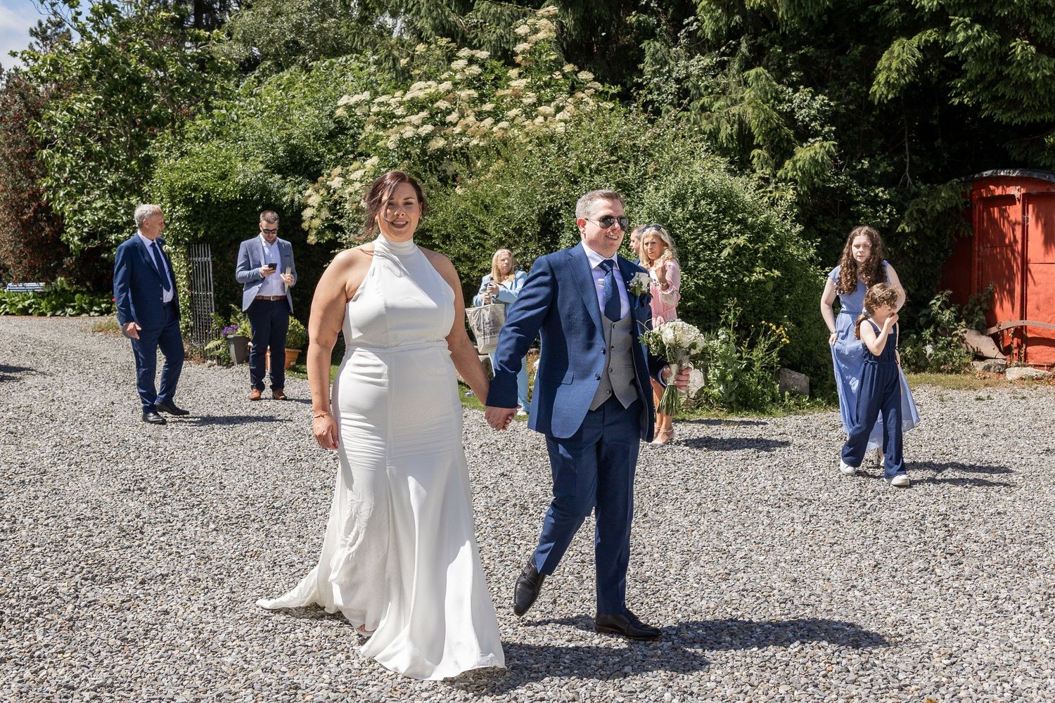 Bride and groom walk hand in hand on gravel path, guests in background, outdoor wedding ceremony on sunny day.
