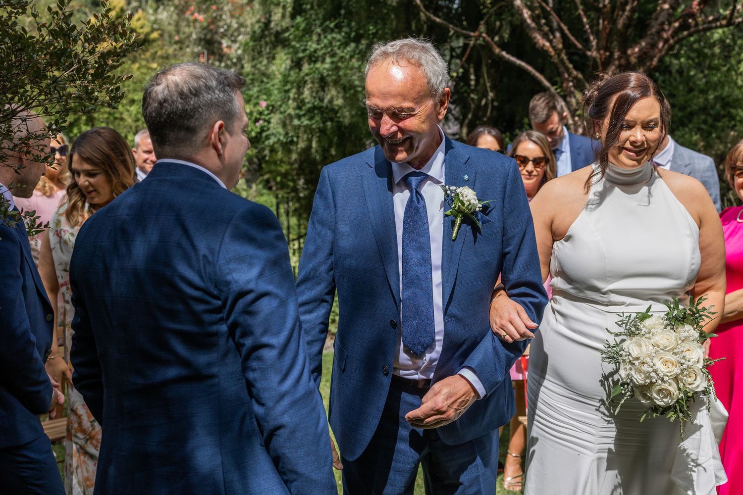 Bride in white halter gown walks with man in blue suit at outdoor wedding ceremony with guests.