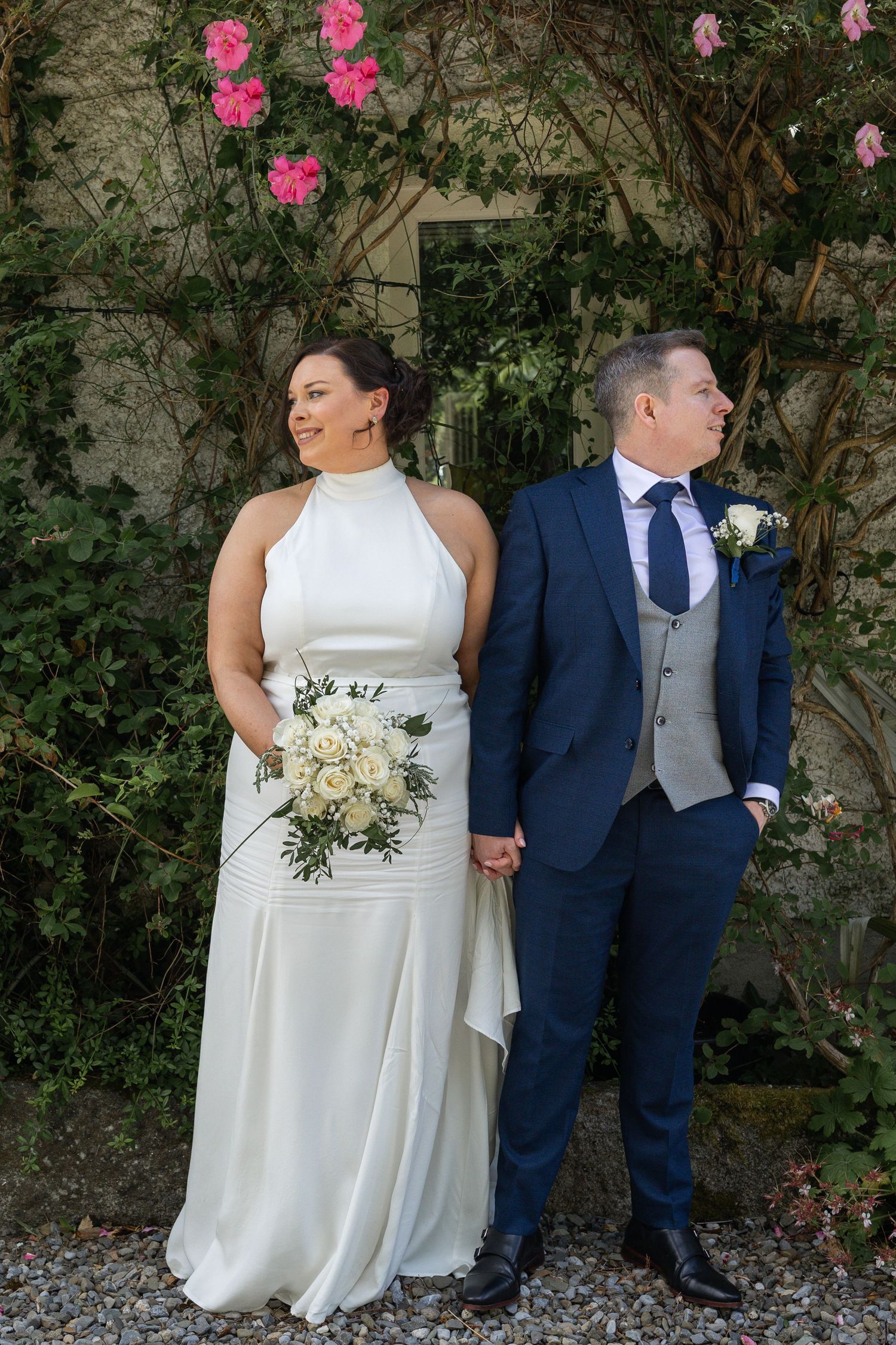Bride in white halter gown and groom in navy suit hold hands before a stone wall with pink roses.