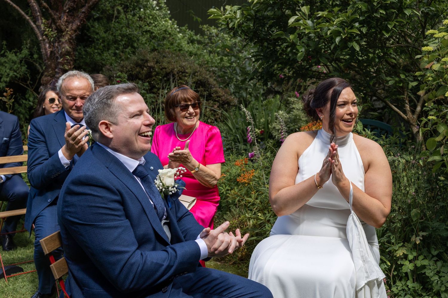 Bride and groom laughing joyfully during outdoor wedding ceremony with guests clapping in sunny garden setting.