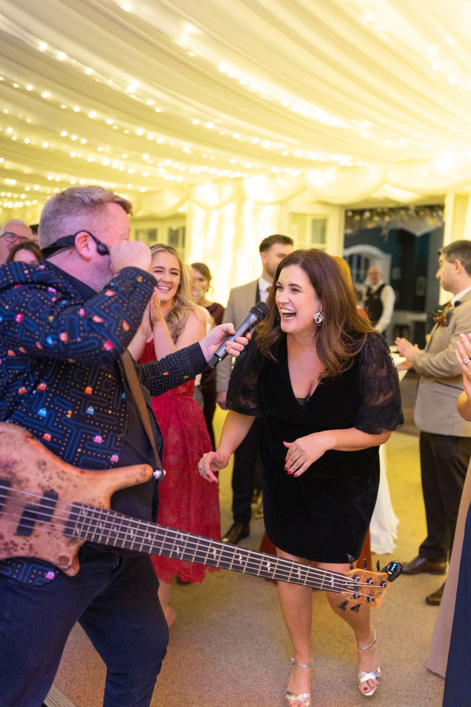Live band performing at a wedding reception, guitarist in colorful shirt holds mic for laughing woman in black dress.