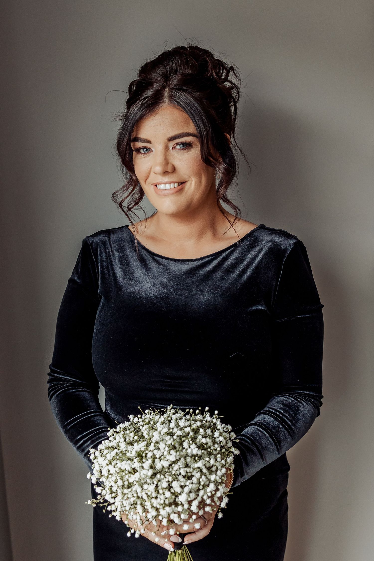 Woman in black velvet dress holding delicate baby's breath bouquet against neutral background.