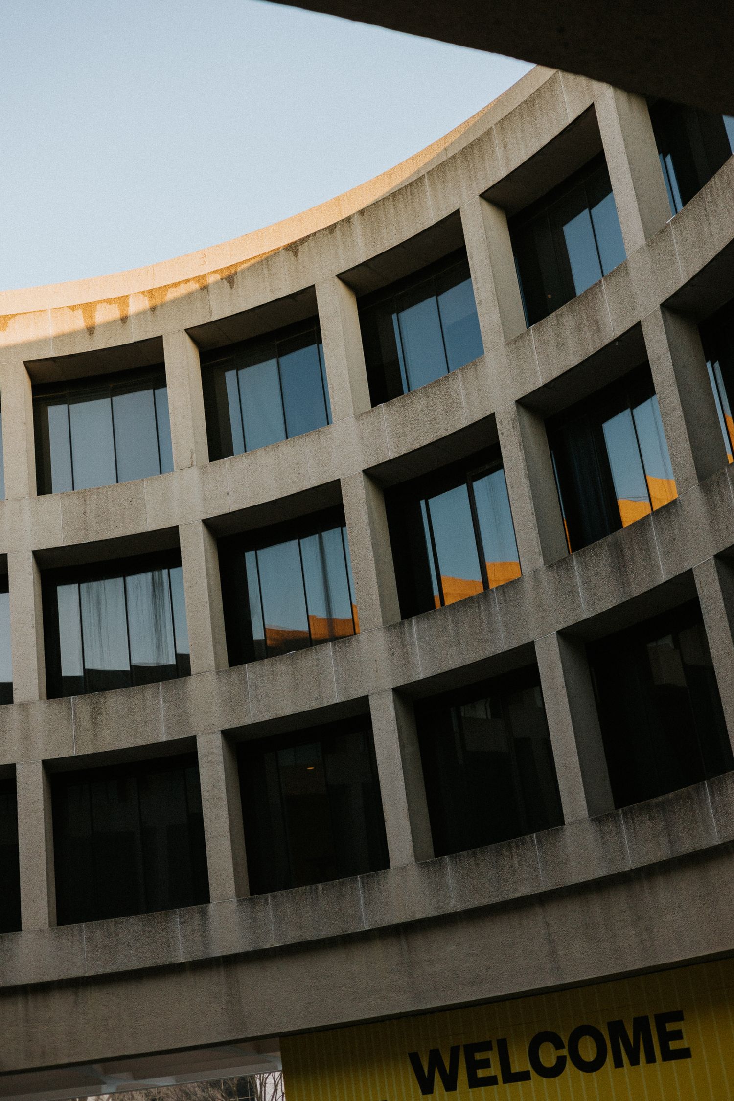 Curved brutalist concrete building facade with grid windows reflecting sky, yellow Welcome sign below.