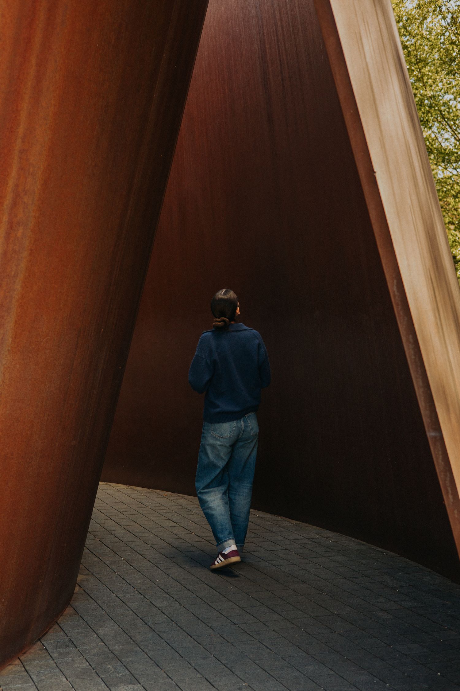 Person in blue hoodie and jeans walking through large curved rusted steel sculpture panels outdoors.
