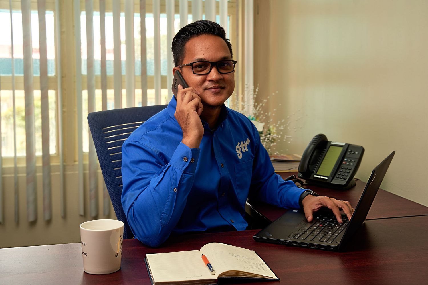 Professional wearing glasses sits at desk with laptop and notebook in sunlit office space.
