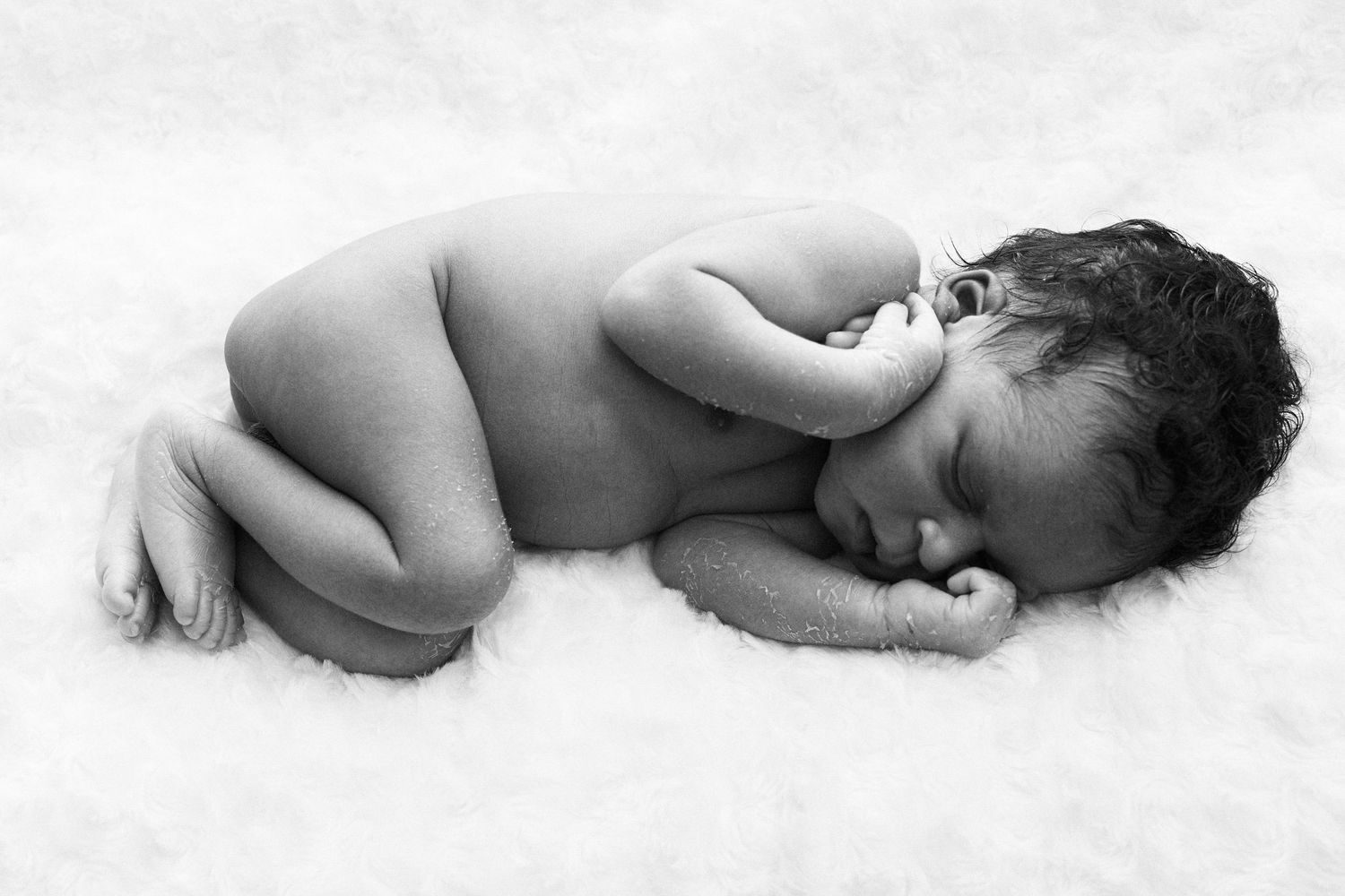 Black and white photo of a sleeping newborn baby curled up on soft white blanket.