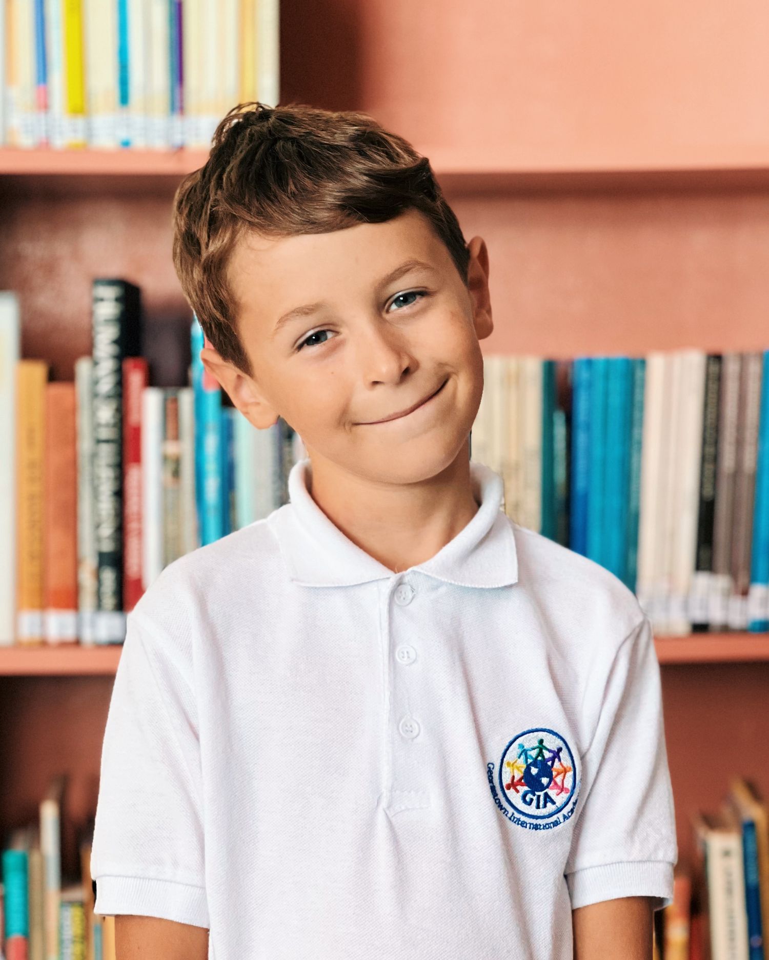 Student in white polo shirt with school emblem smiles warmly in library setting with bookshelves in background.
