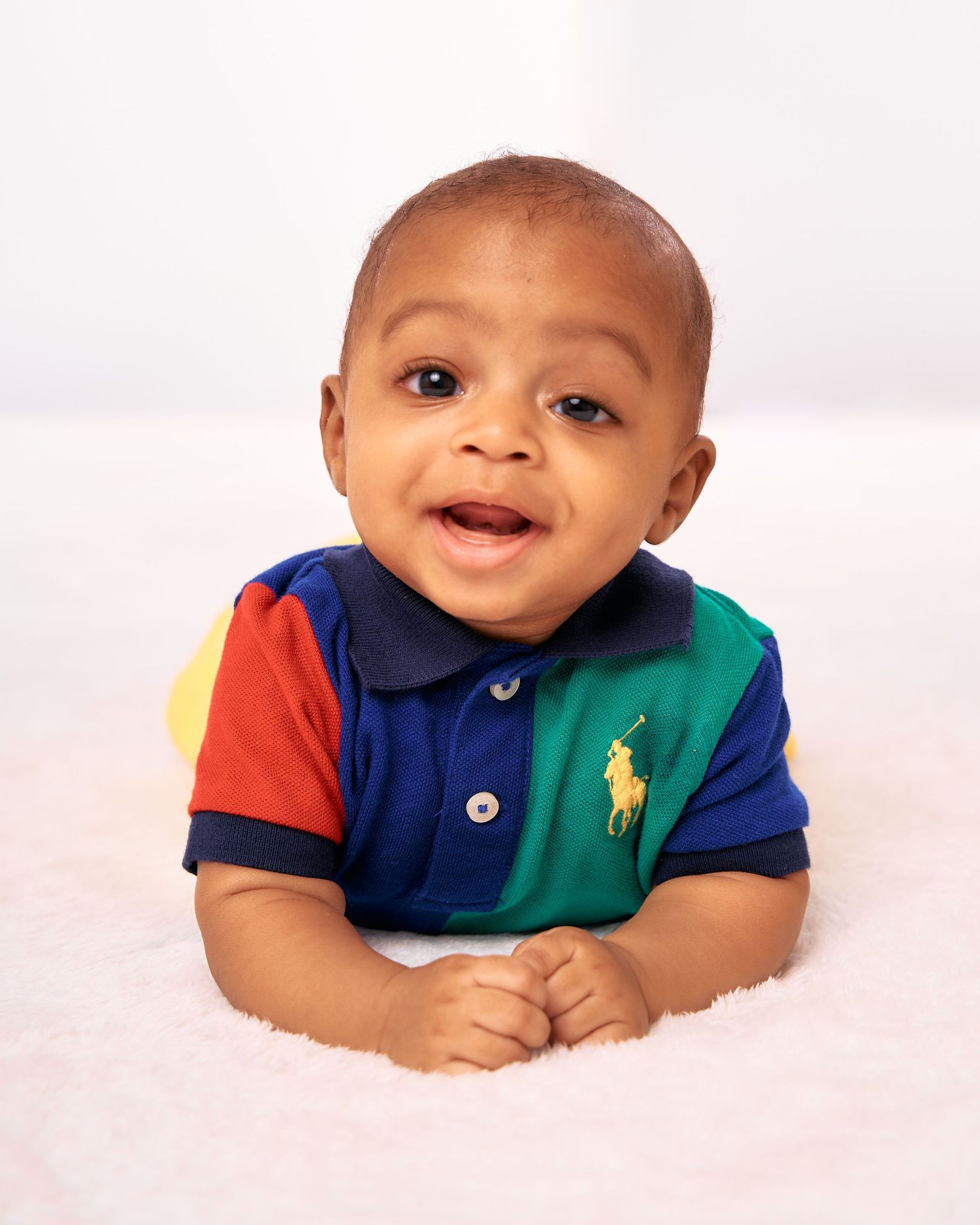 A baby wearing a colorful polo shirt smiles while lying on their stomach against a white background.