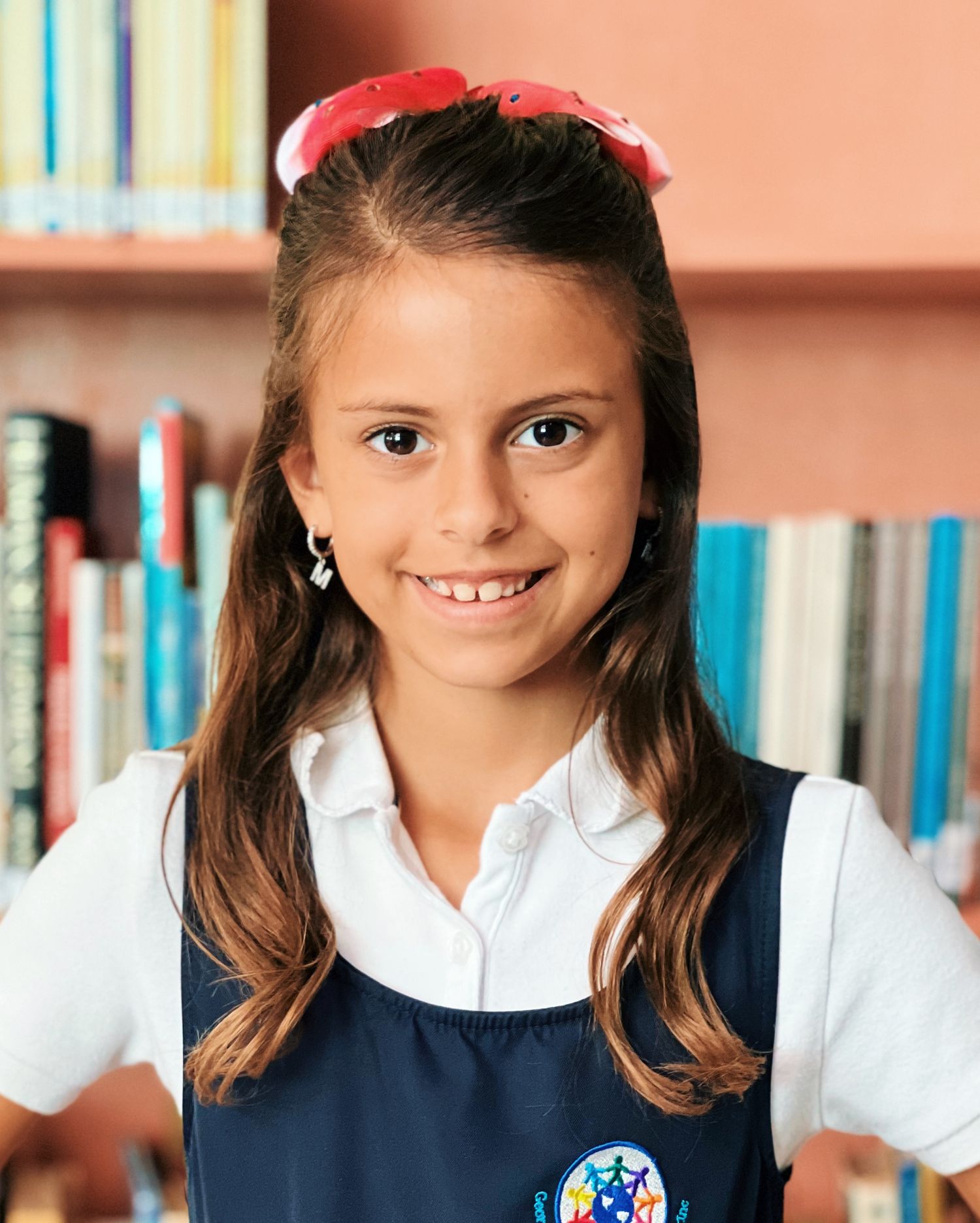 Young student in navy blue uniform and white collared shirt smiles brightly in school library setting.