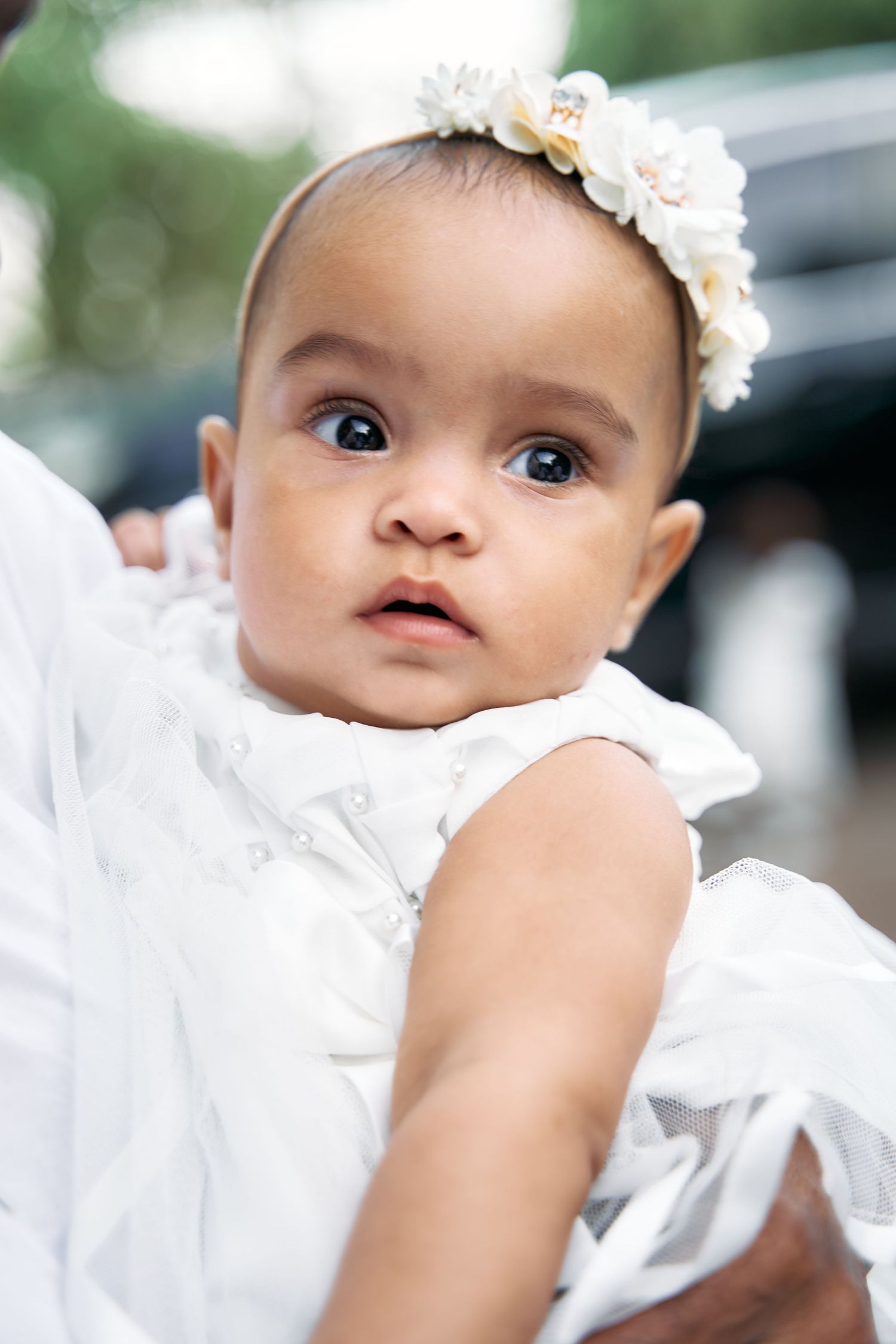 Baby wearing white dress and floral headband looks up with curious expression during outdoor portrait session.