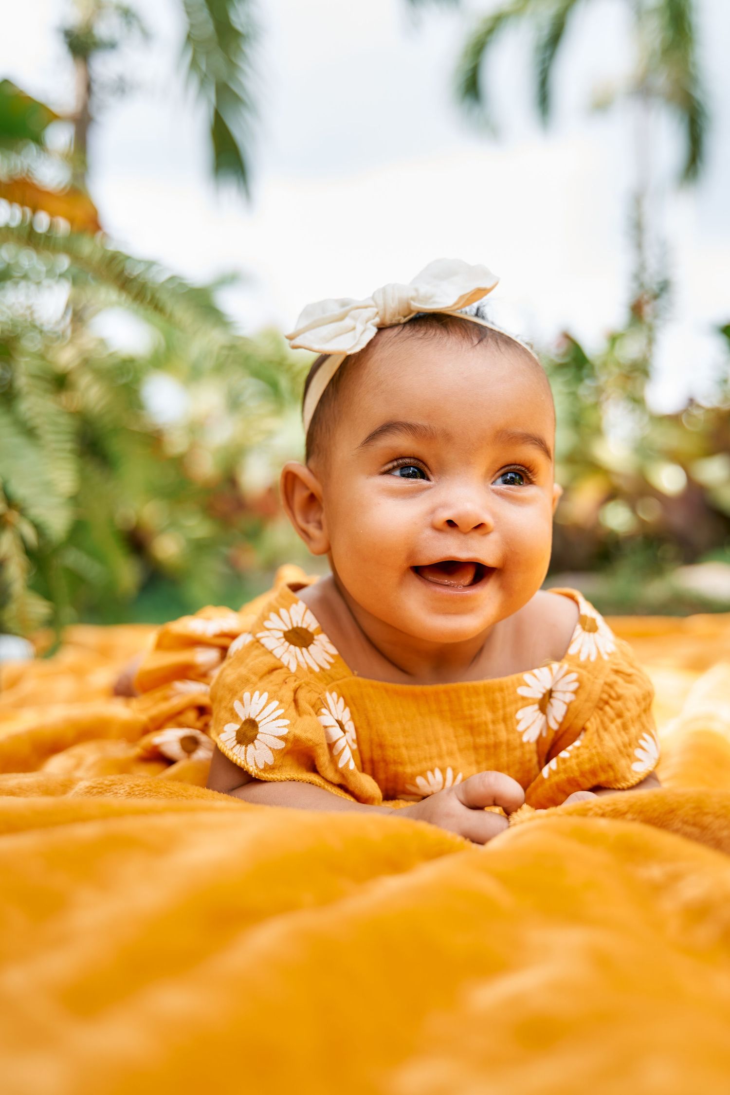 Adorable baby in yellow outfit lying on orange blanket with palm trees in background smiling joyfully at camera.