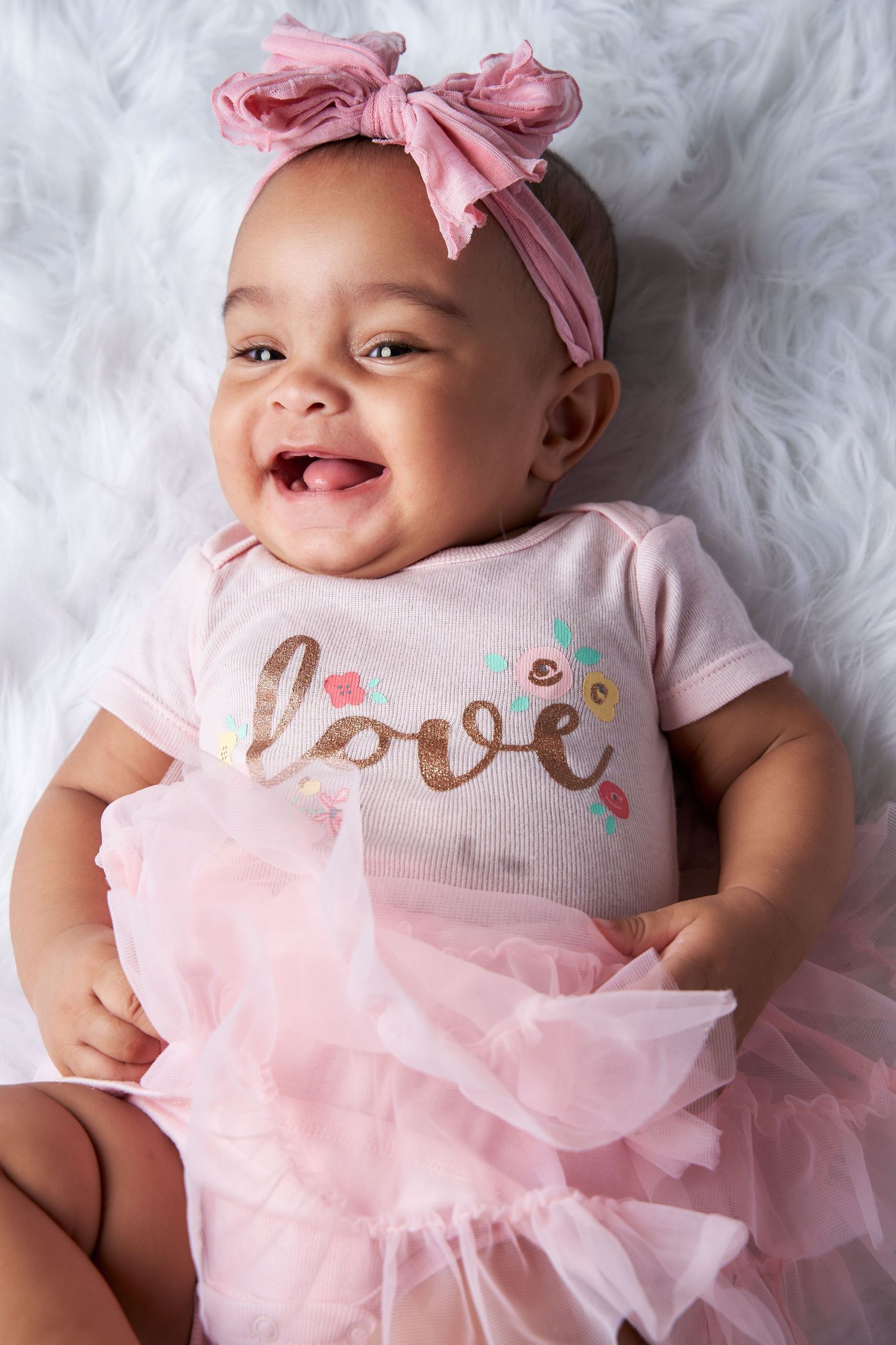 Baby wearing pink tulle dress and bow headband lies on white fluffy blanket.