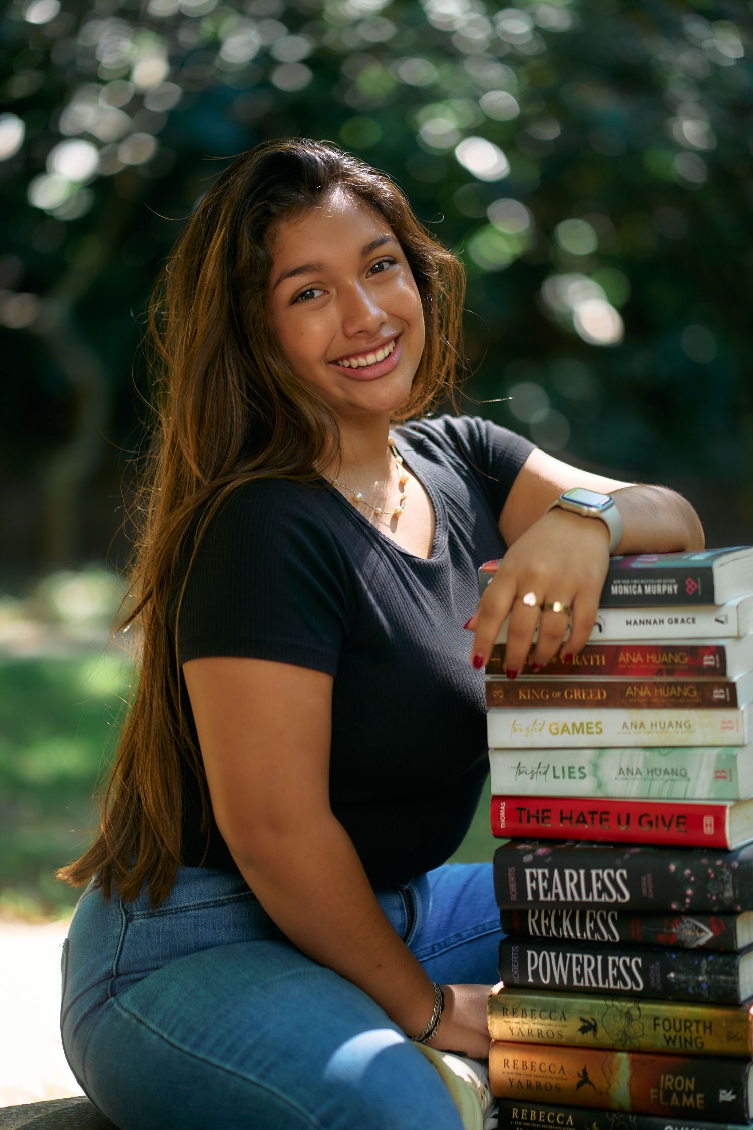 A person poses outdoors with a stack of books in casual attire with natural bokeh background.