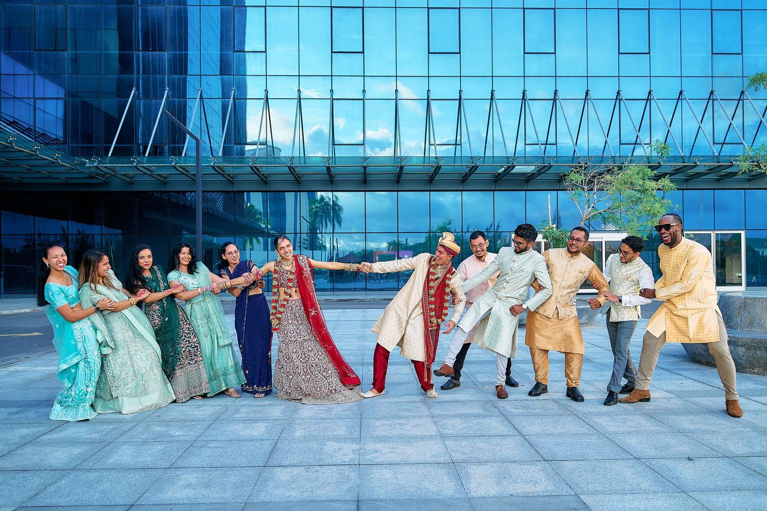 Panoramic shot of wedding party in turquoise and colorful outfits lined up against glass building.