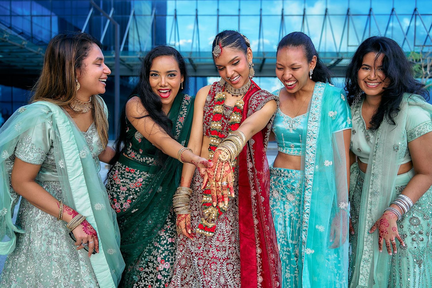 Wedding party in colorful Indian attire poses playfully against modern glass building.
