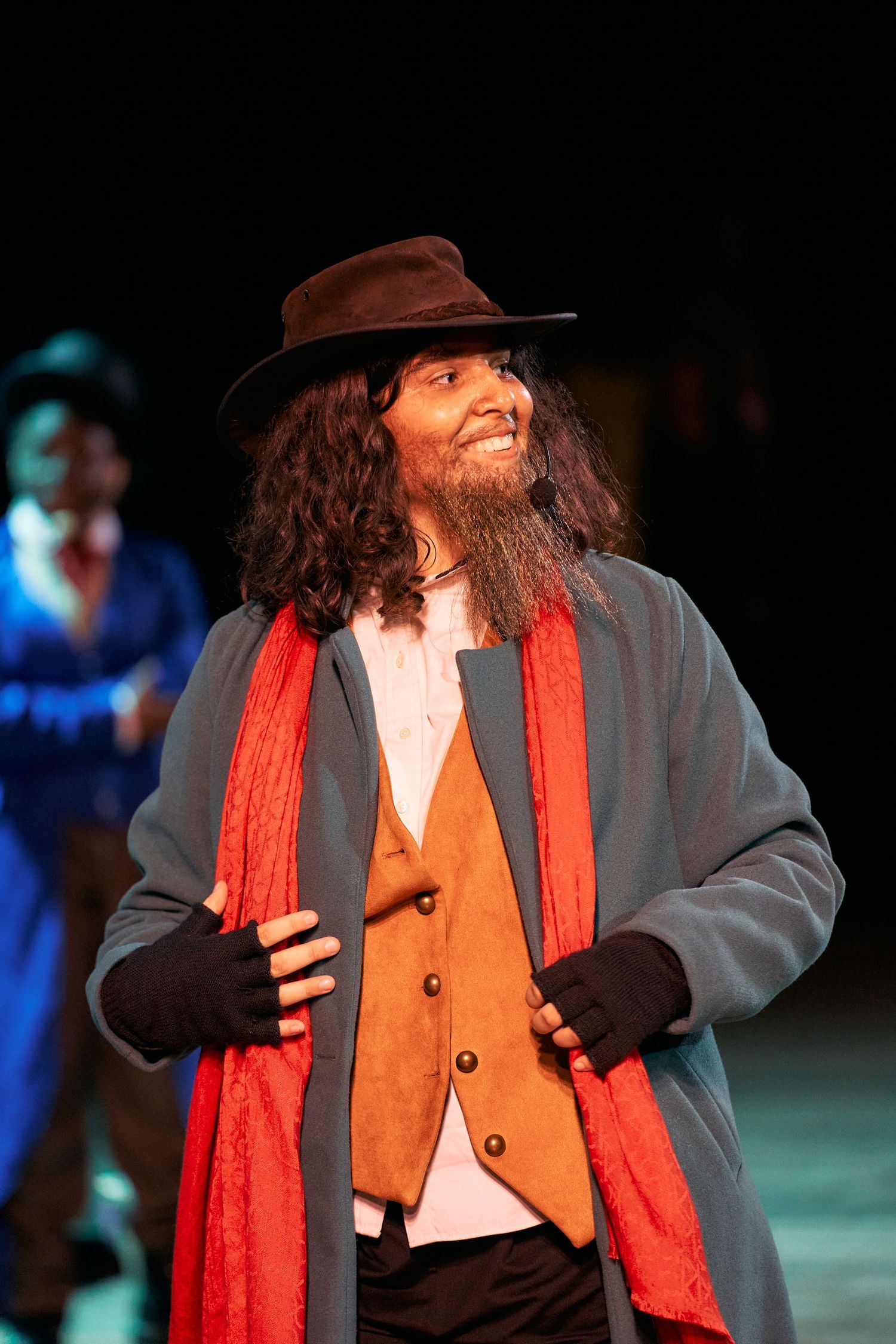 A performer in a brown hat, red scarf, and gray coat smiles during a theatrical production on stage.