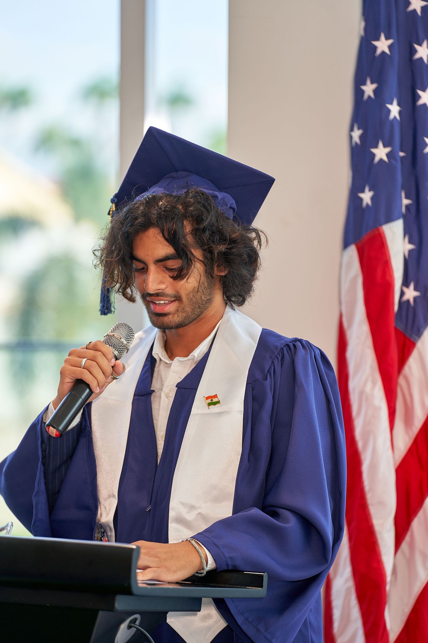 Graduate in blue cap and gown gives speech at podium with American flag in background.