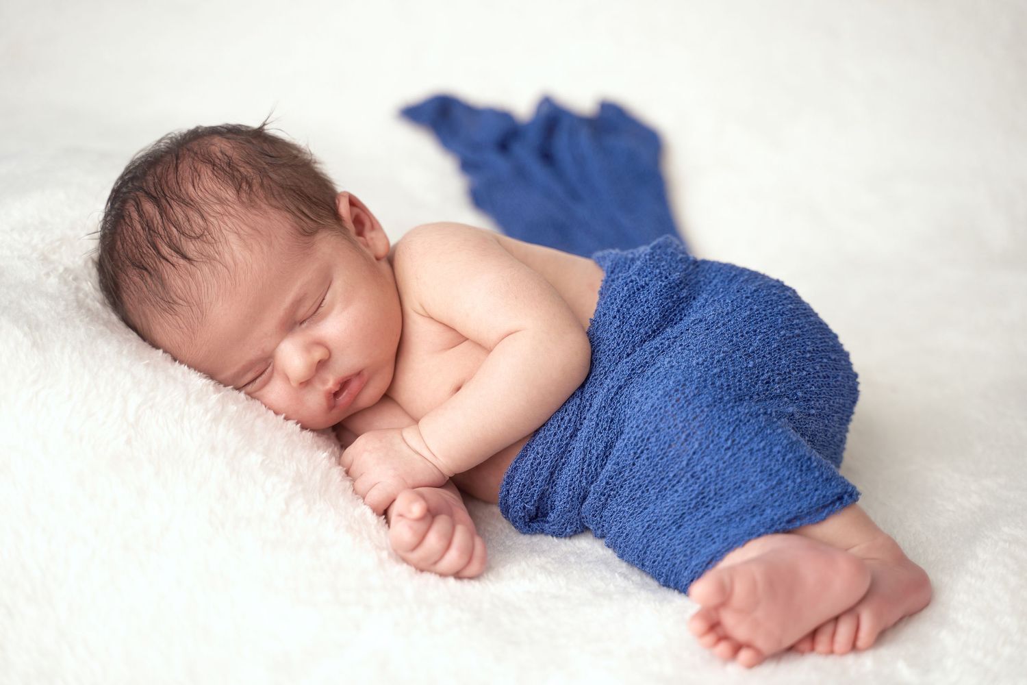 Sleeping newborn baby wrapped in soft blue fabric lying on white blanket in peaceful pose.