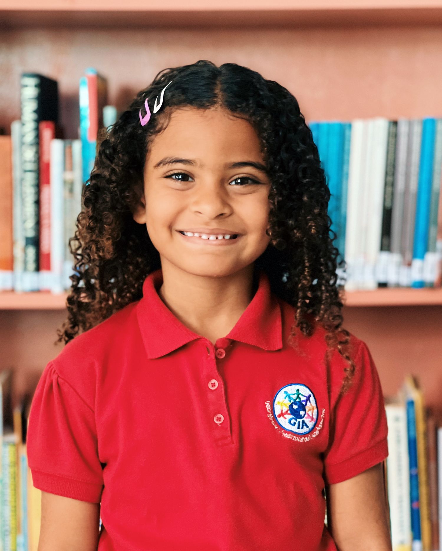 Student in red polo uniform with curly hair smiling at camera against bookshelf background.