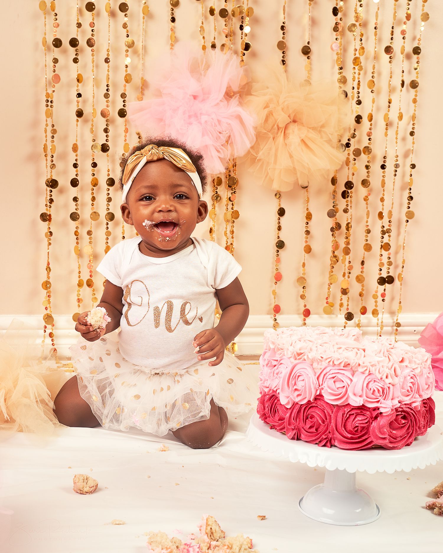 Baby wearing white dress and bow smiles during first birthday cake smash photo session with pink floral backdrop.