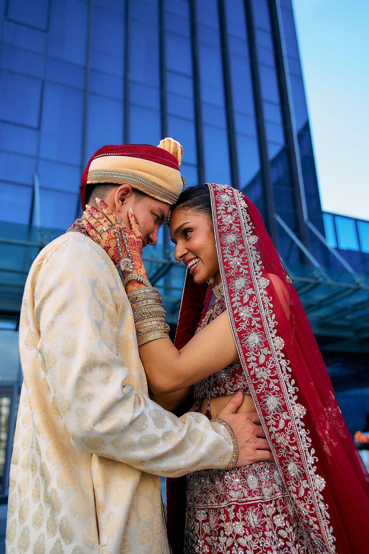 Tender moments captured between newlyweds in traditional Indian wedding dress against modern architecture.