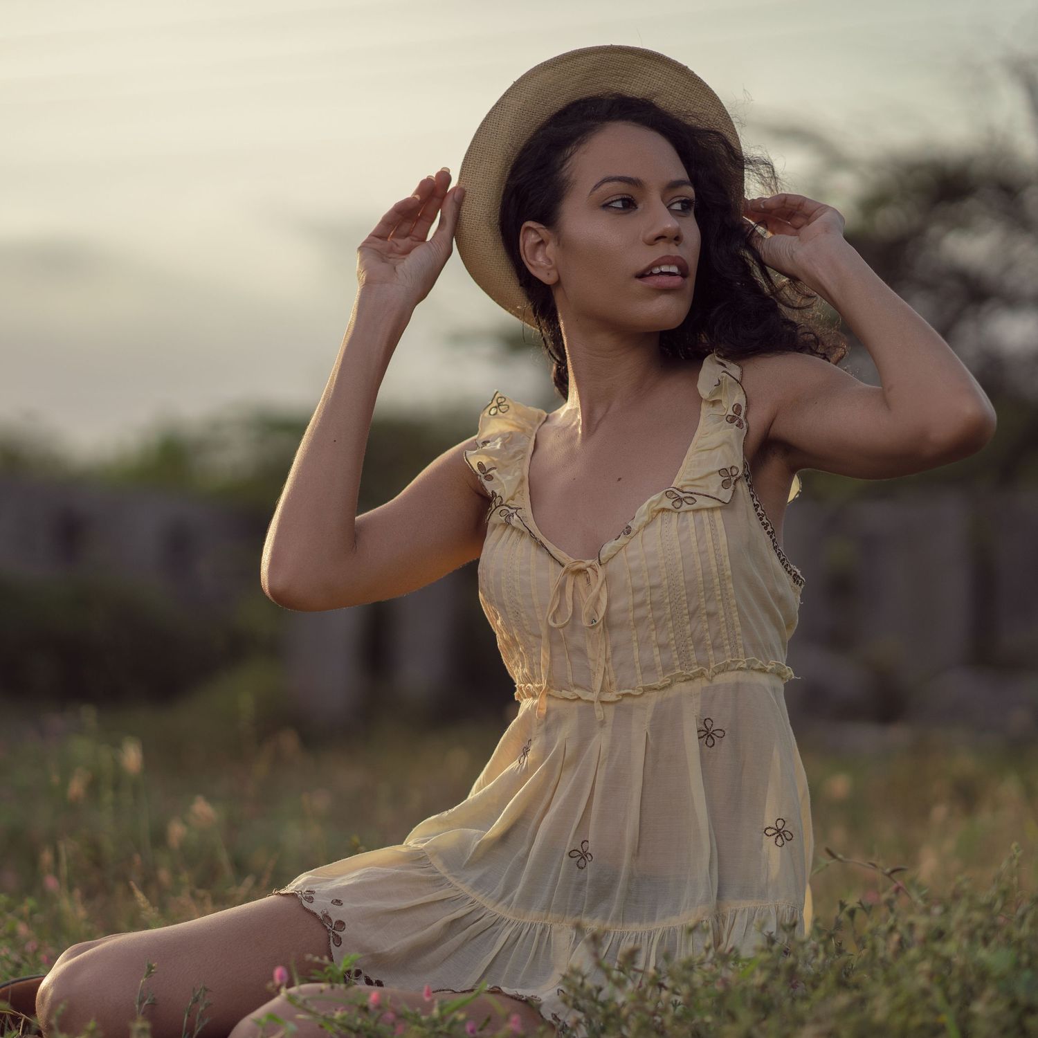 Dreamy portrait of a person in a cream dress and brown hat in a field at dusk.