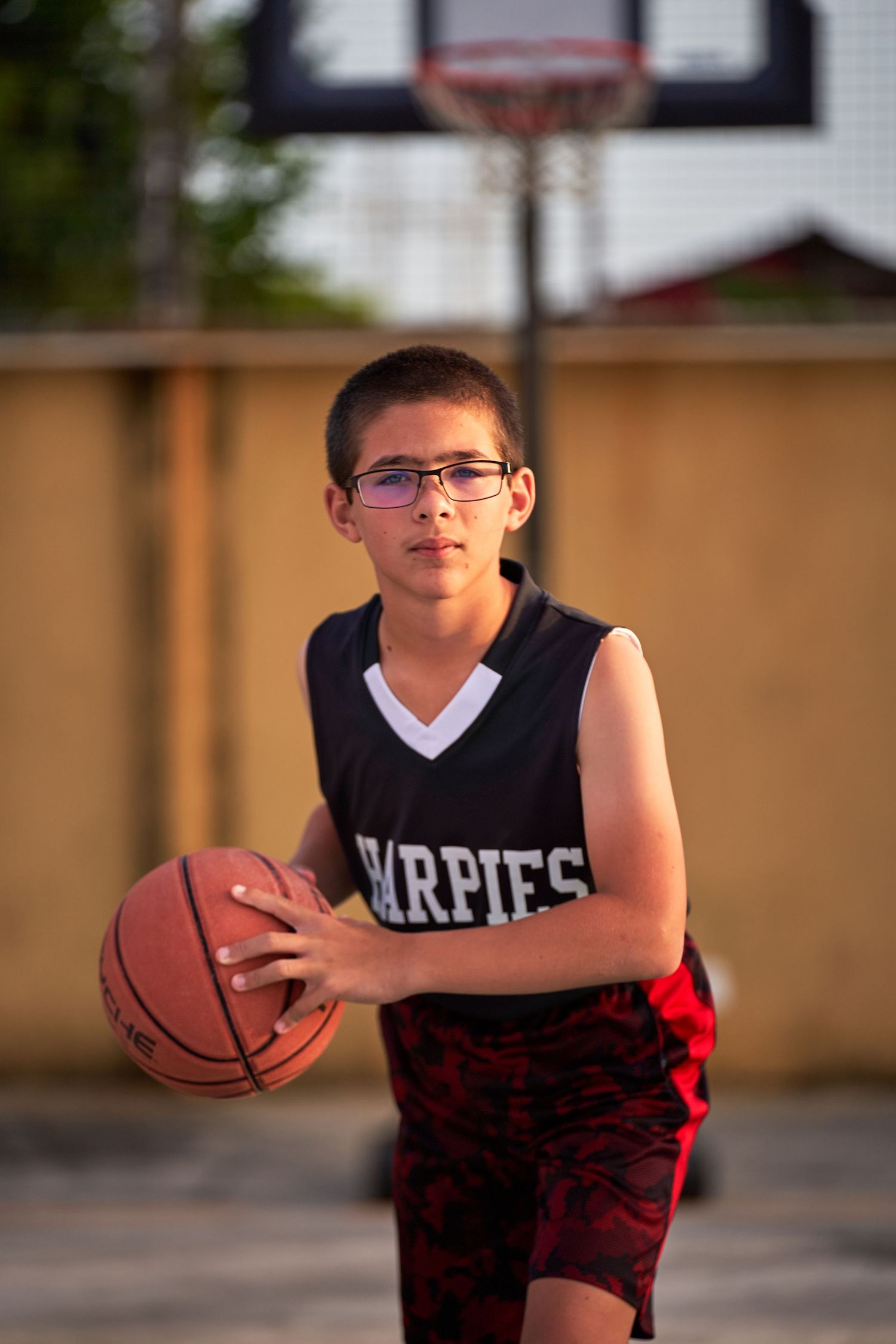 A young basketball player in a red and black uniform practices dribbling moves on an outdoor court.