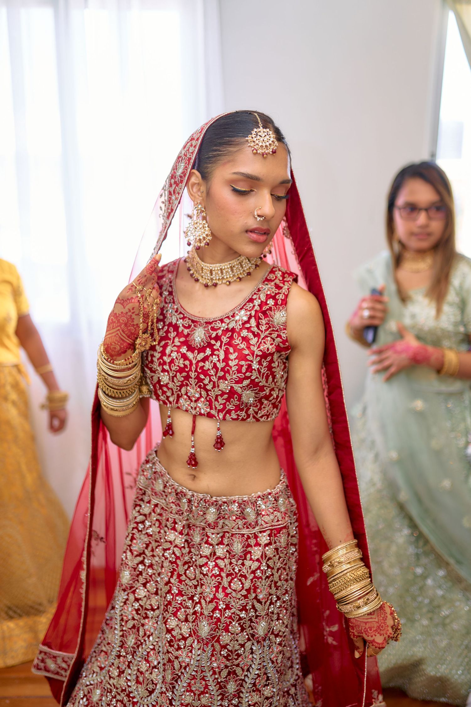 Indian bride in red embroidered lehenga getting ready with bridesmaids in background.