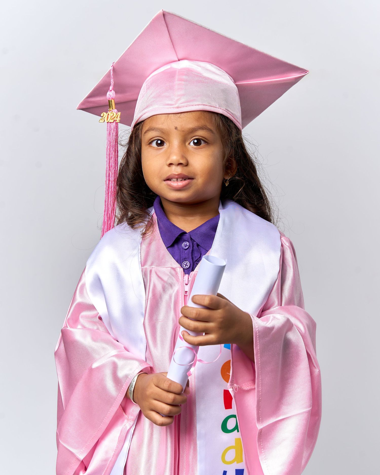 Graduate in pink cap and gown holding diploma during graduation photo session against white background.