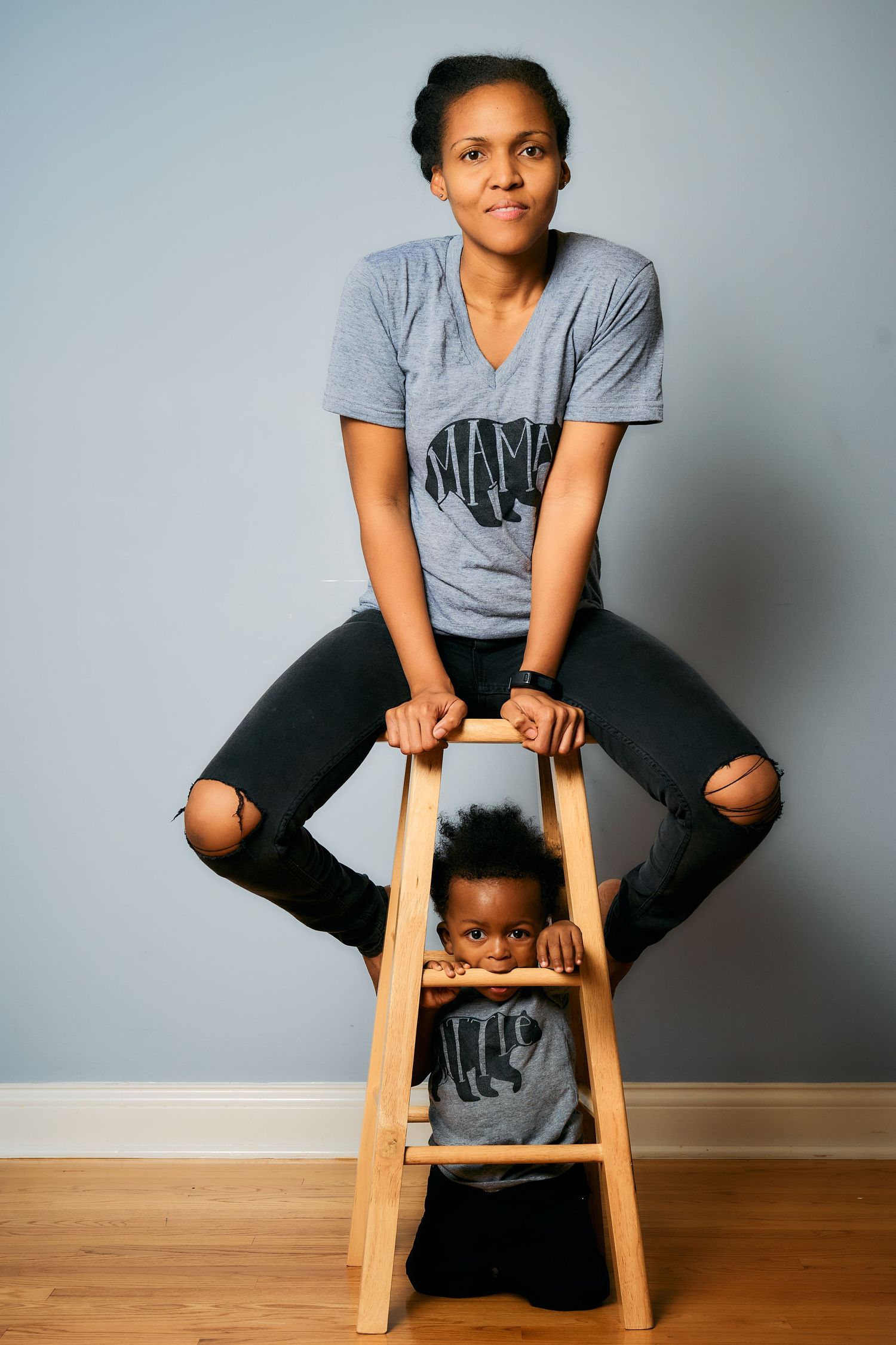 A person in a gray t-shirt and ripped jeans sits casually on a wooden stool against a light gray backdrop.