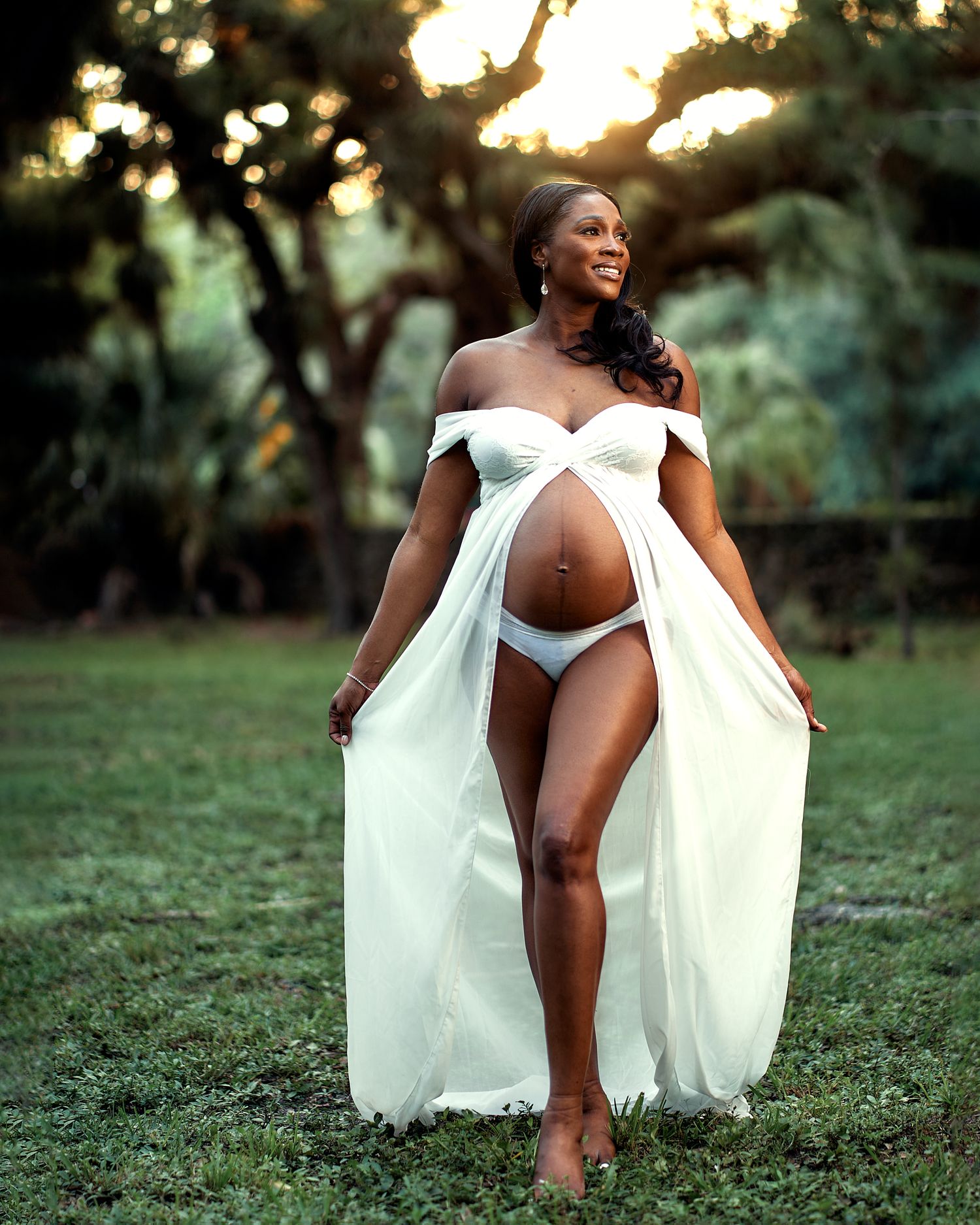 Maternity photo shoot in flowing white dress on grassy field at sunset.