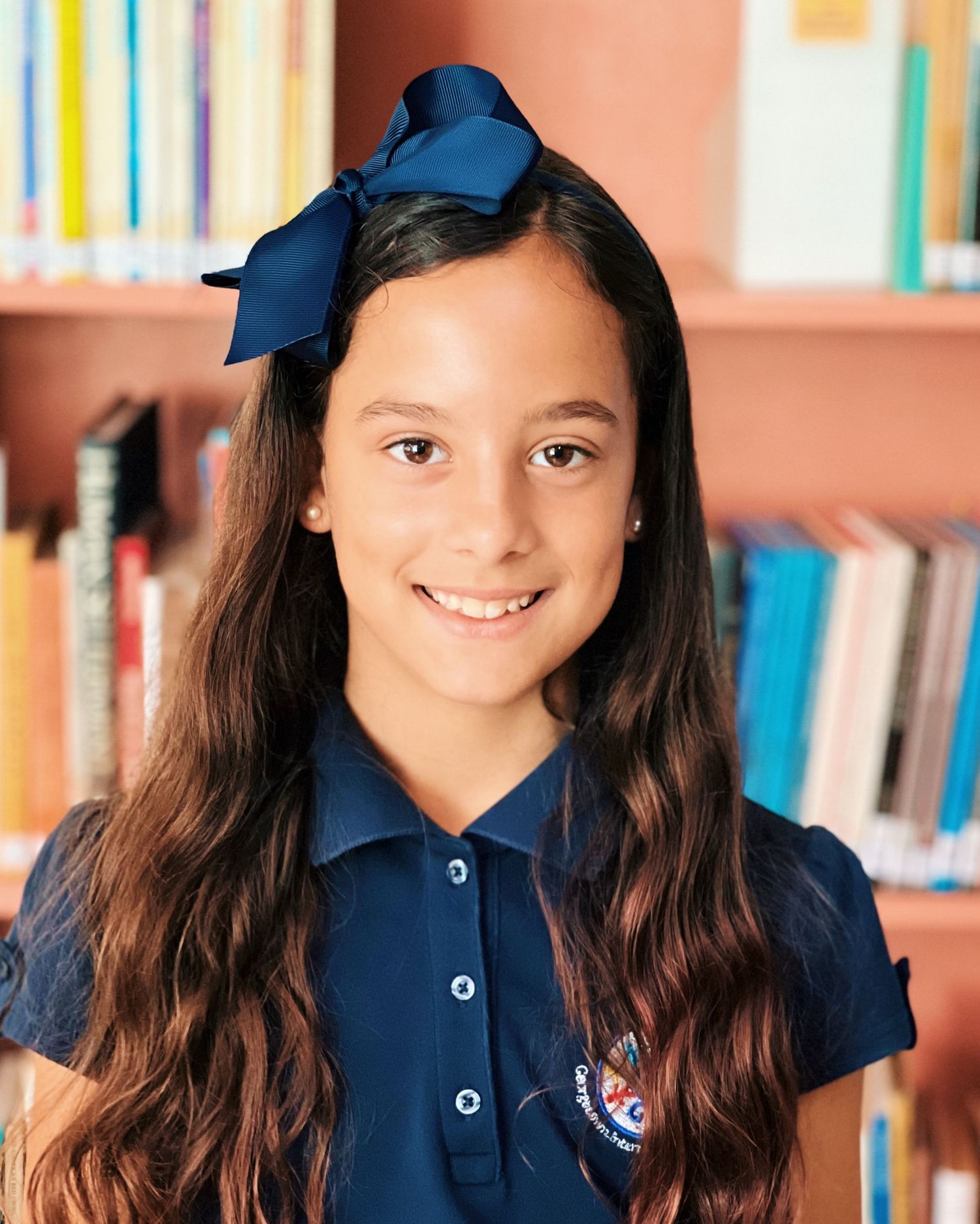 Student in navy blue school uniform smiles while standing in front of colorful library bookshelves.