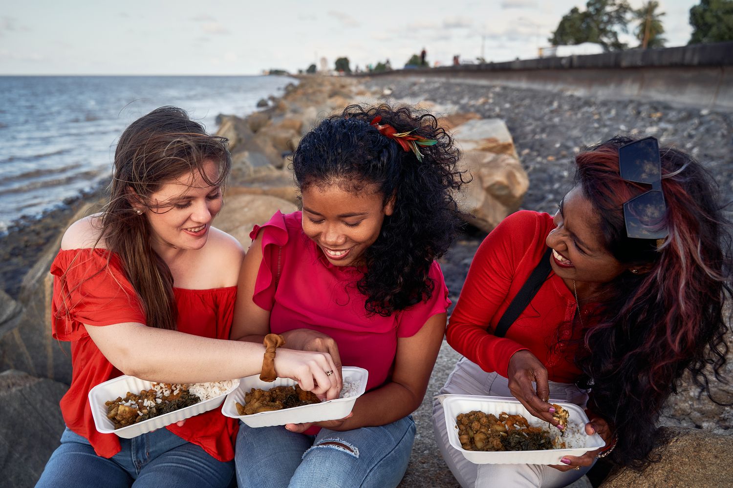 Friends in red tops sharing food and laughing together by the ocean.
