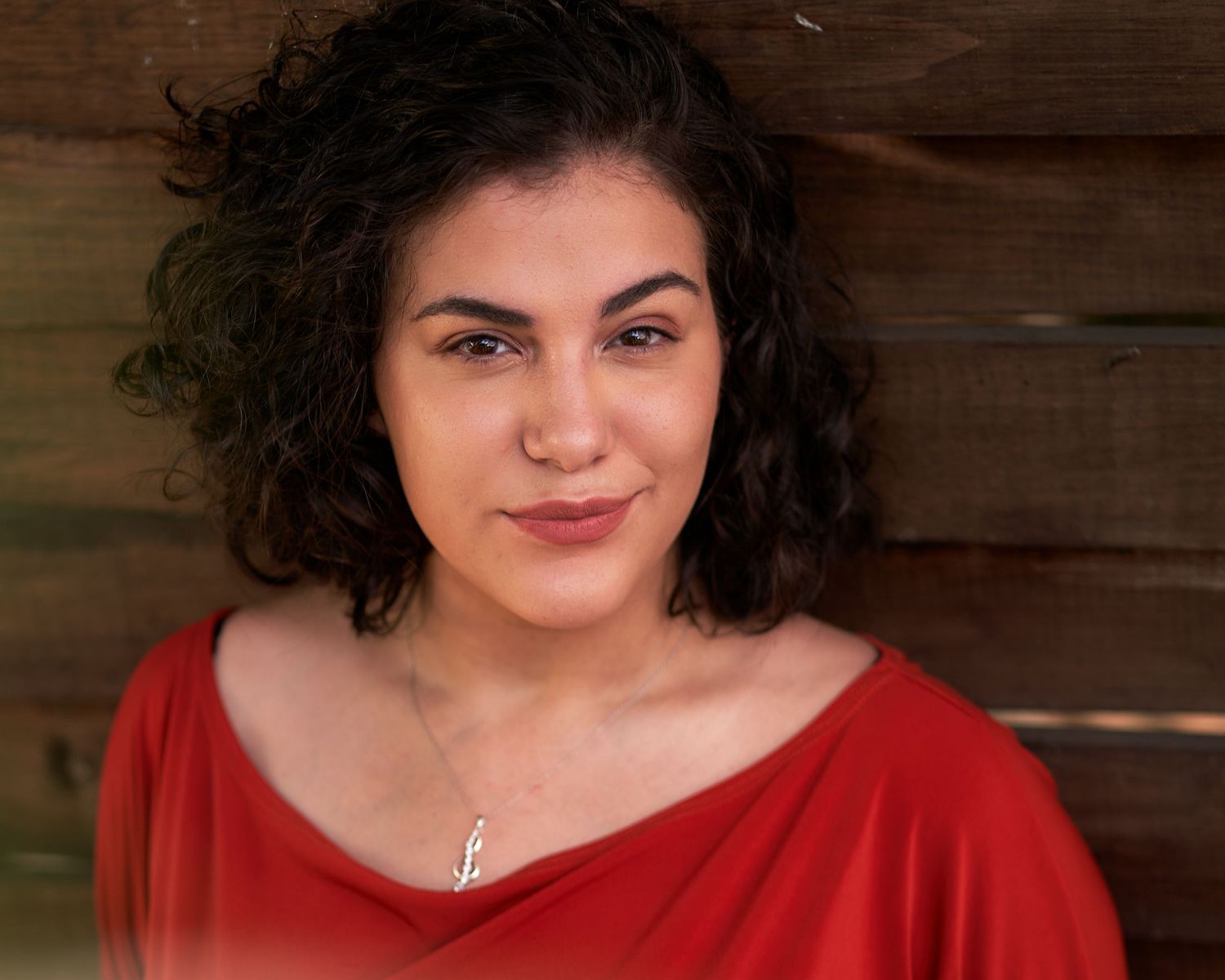 Portrait of a person with curly dark hair wearing a red top against a wooden background.