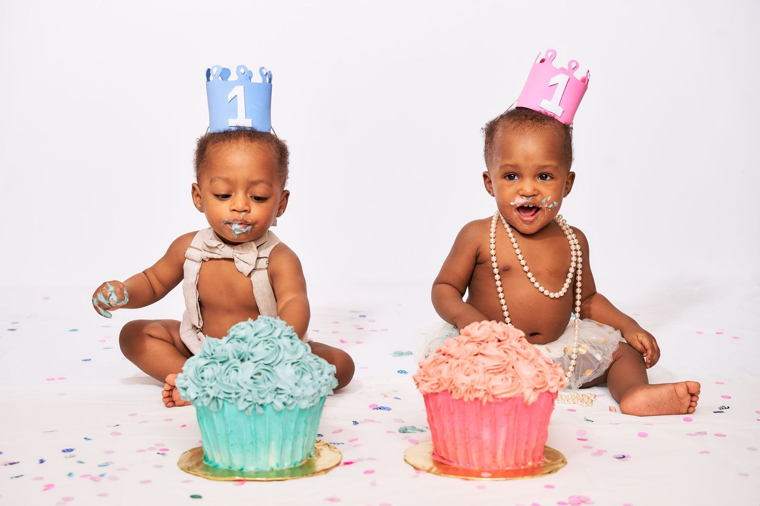 Two babies wearing party attire sit next to colorful cupcakes with confetti during a cake smash photo session.