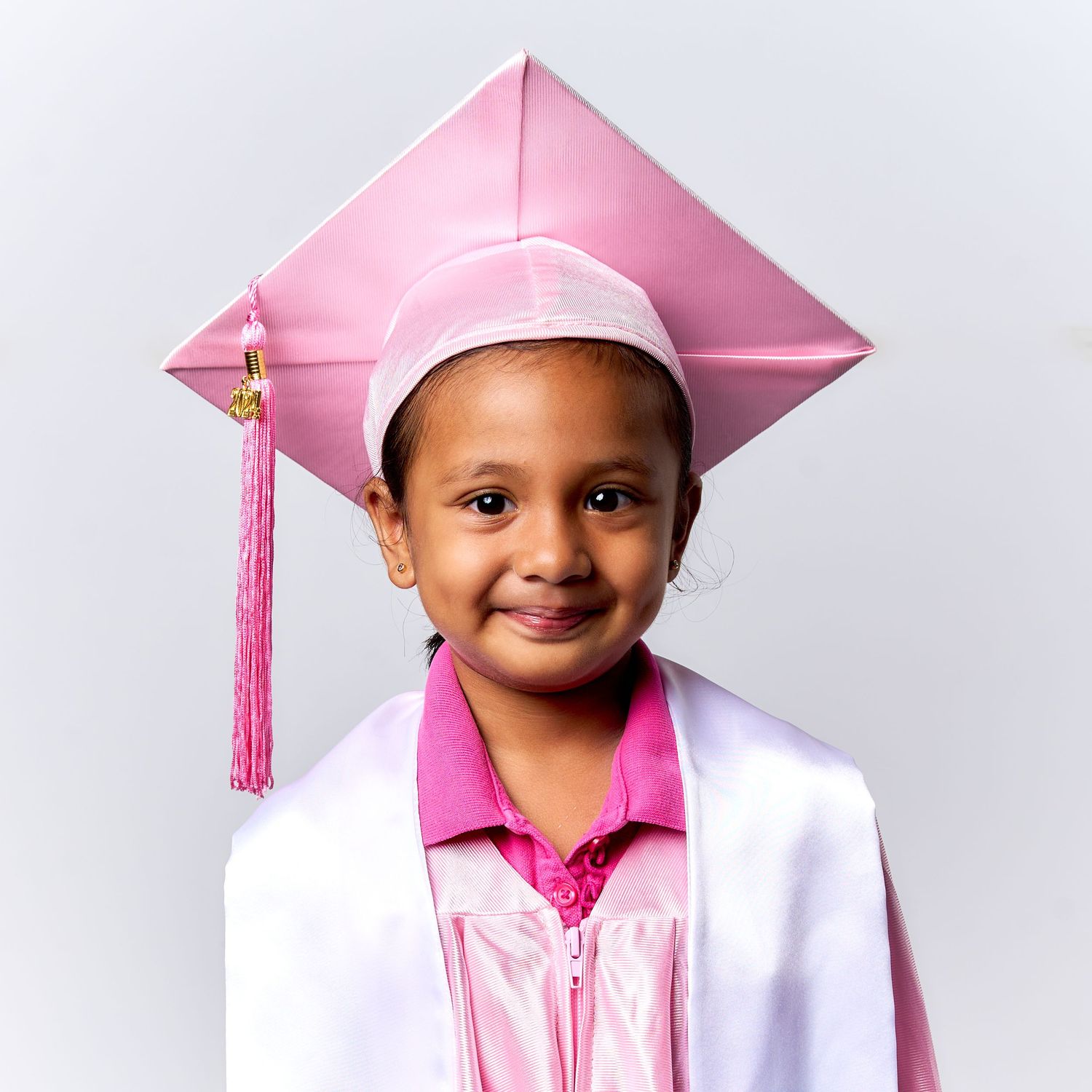 Young graduate wearing pink cap and gown with white jacket poses against light background.