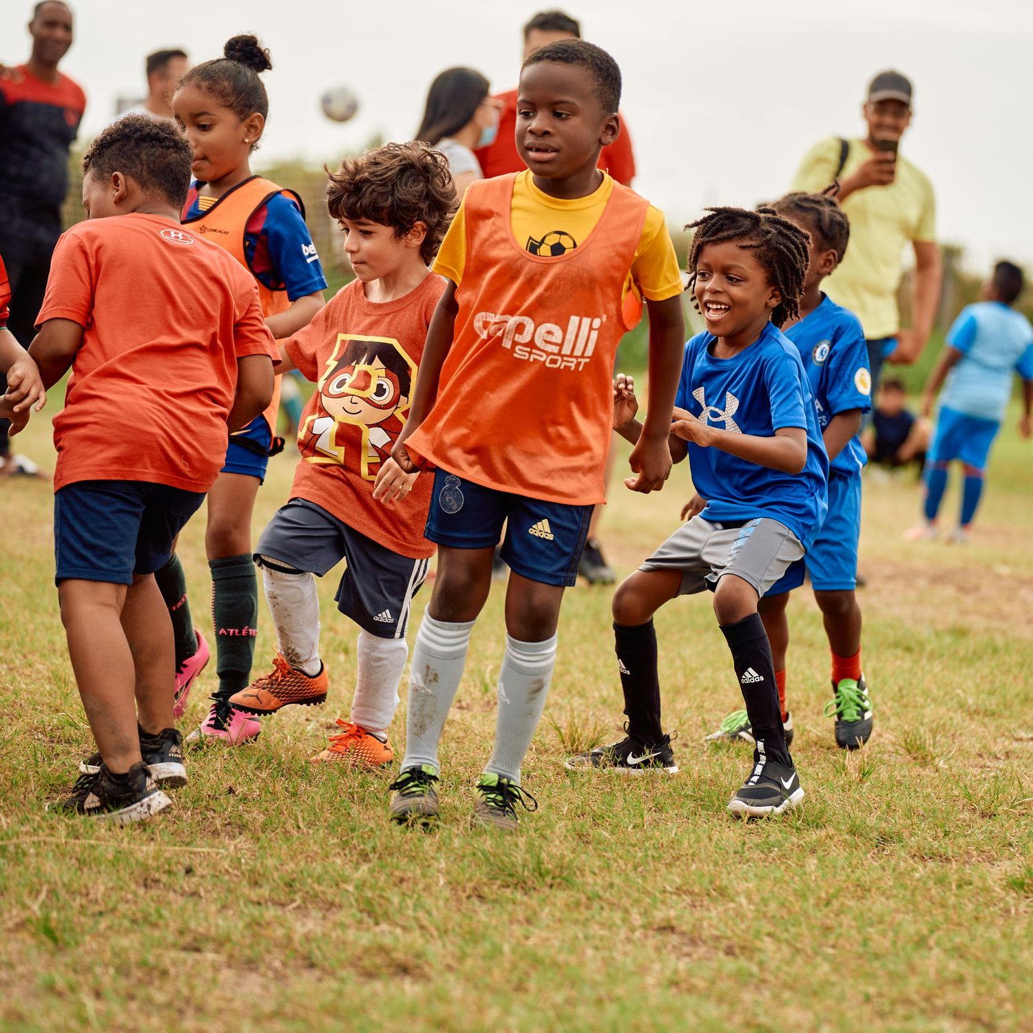 Young soccer players in orange and blue jerseys running and playing together on a grassy field during practice.