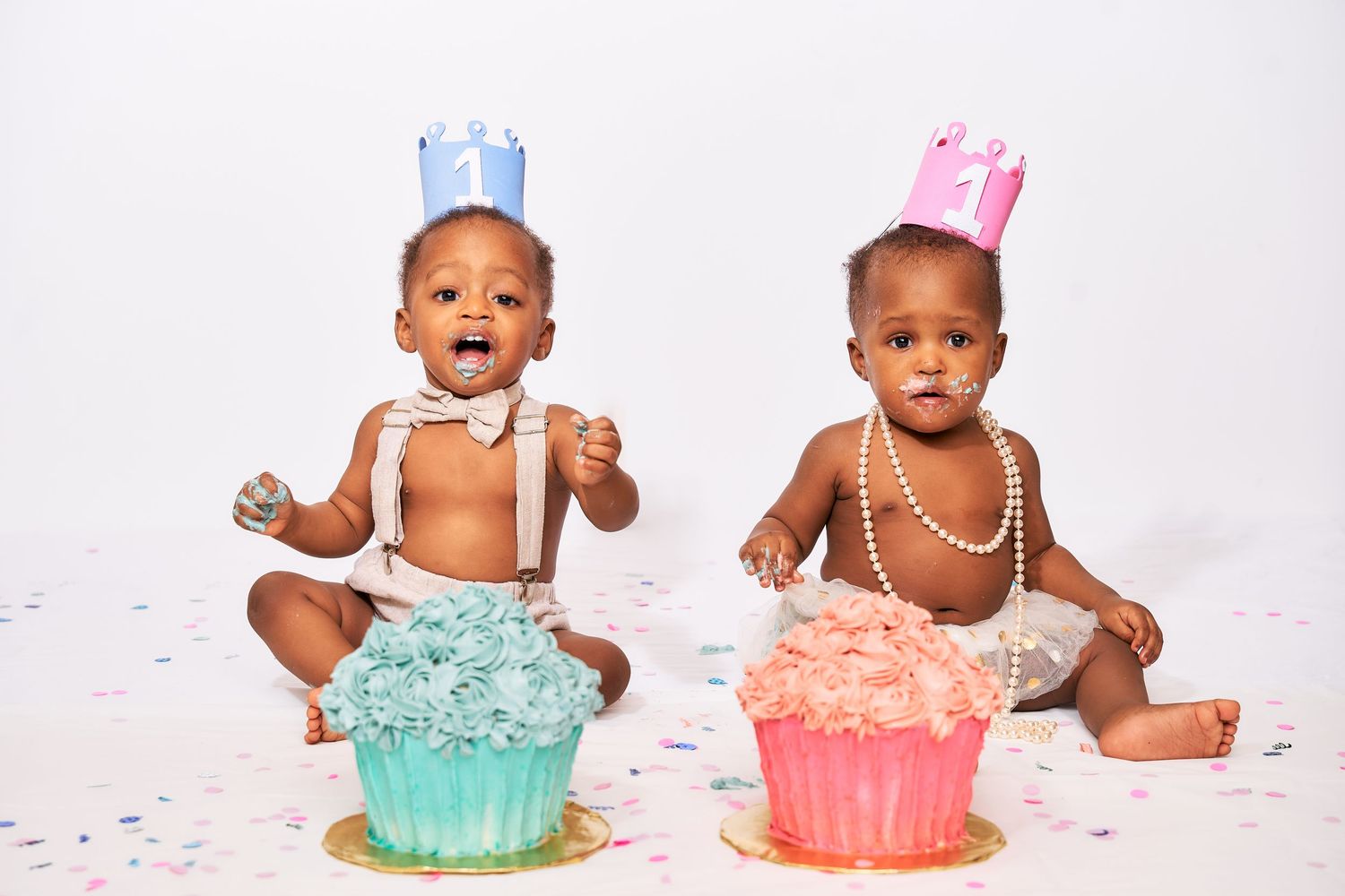 Two babies wearing crowns and pearls sit with giant cupcakes during a first birthday cake smash photoshoot.