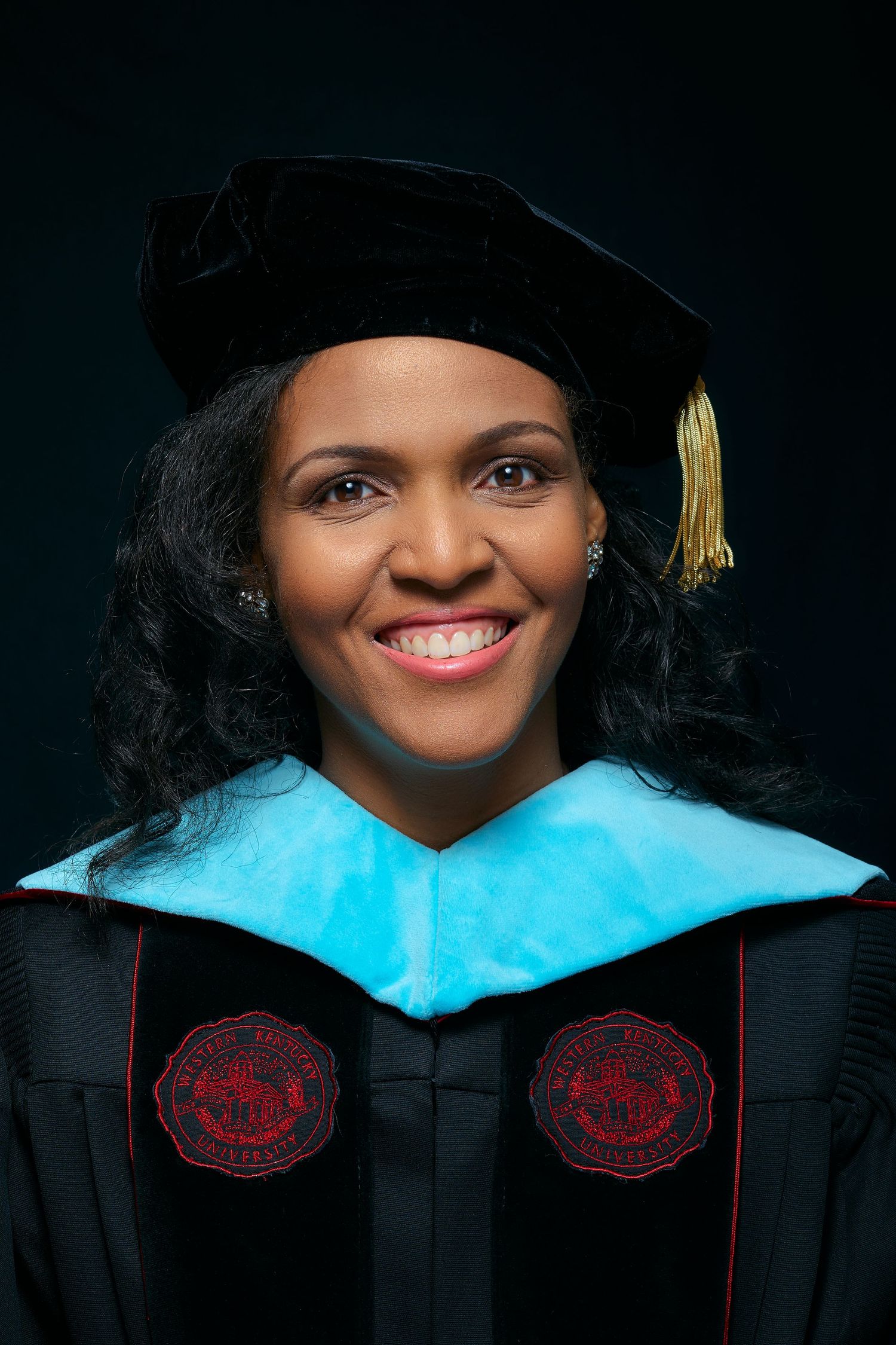 Graduate in academic regalia wearing doctoral tam and gown with blue hood and red emblems smiles warmly in formal portrait.