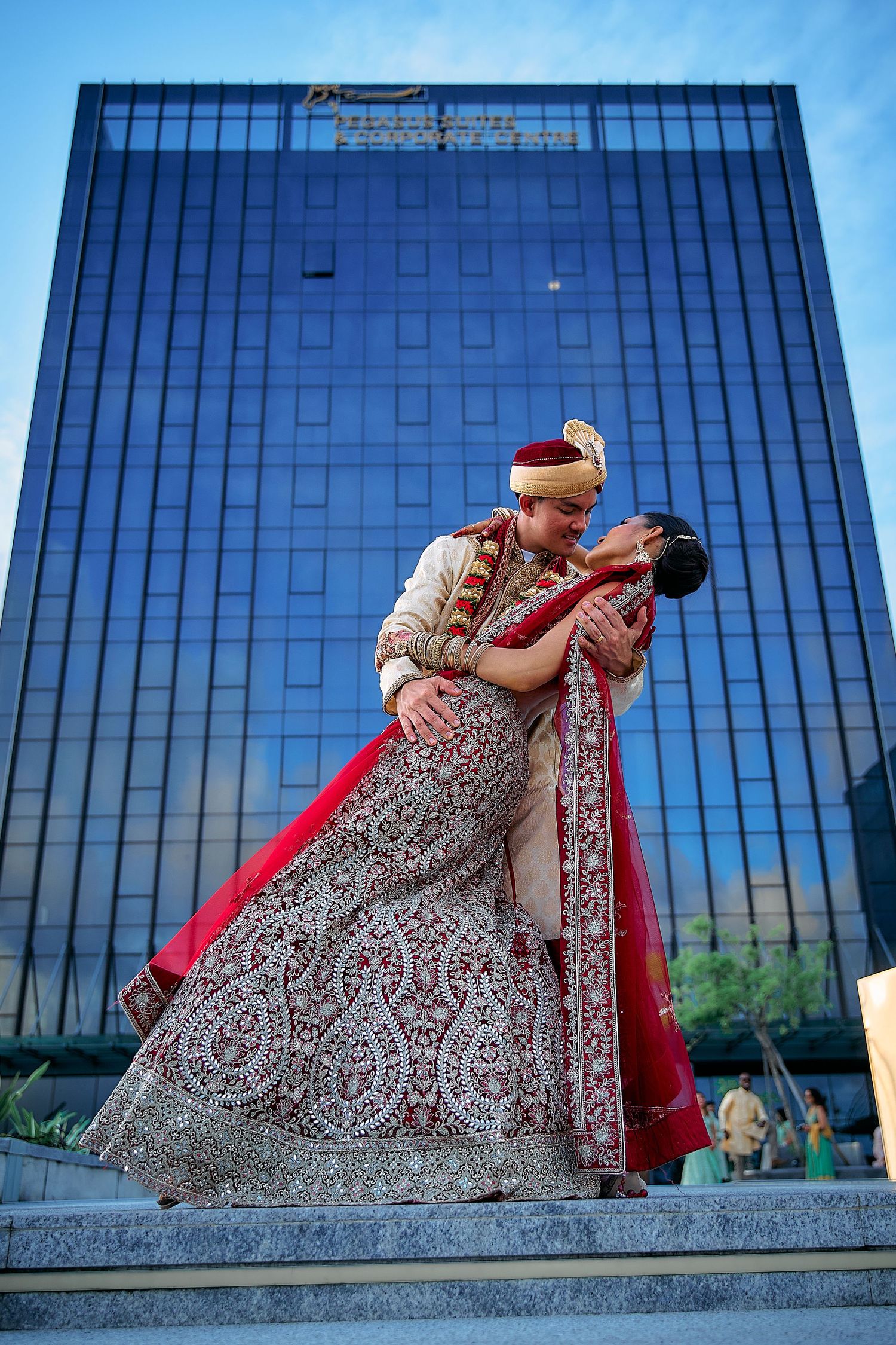 A romantic couple in traditional Indian wedding attire poses dramatically against a modern glass building.
