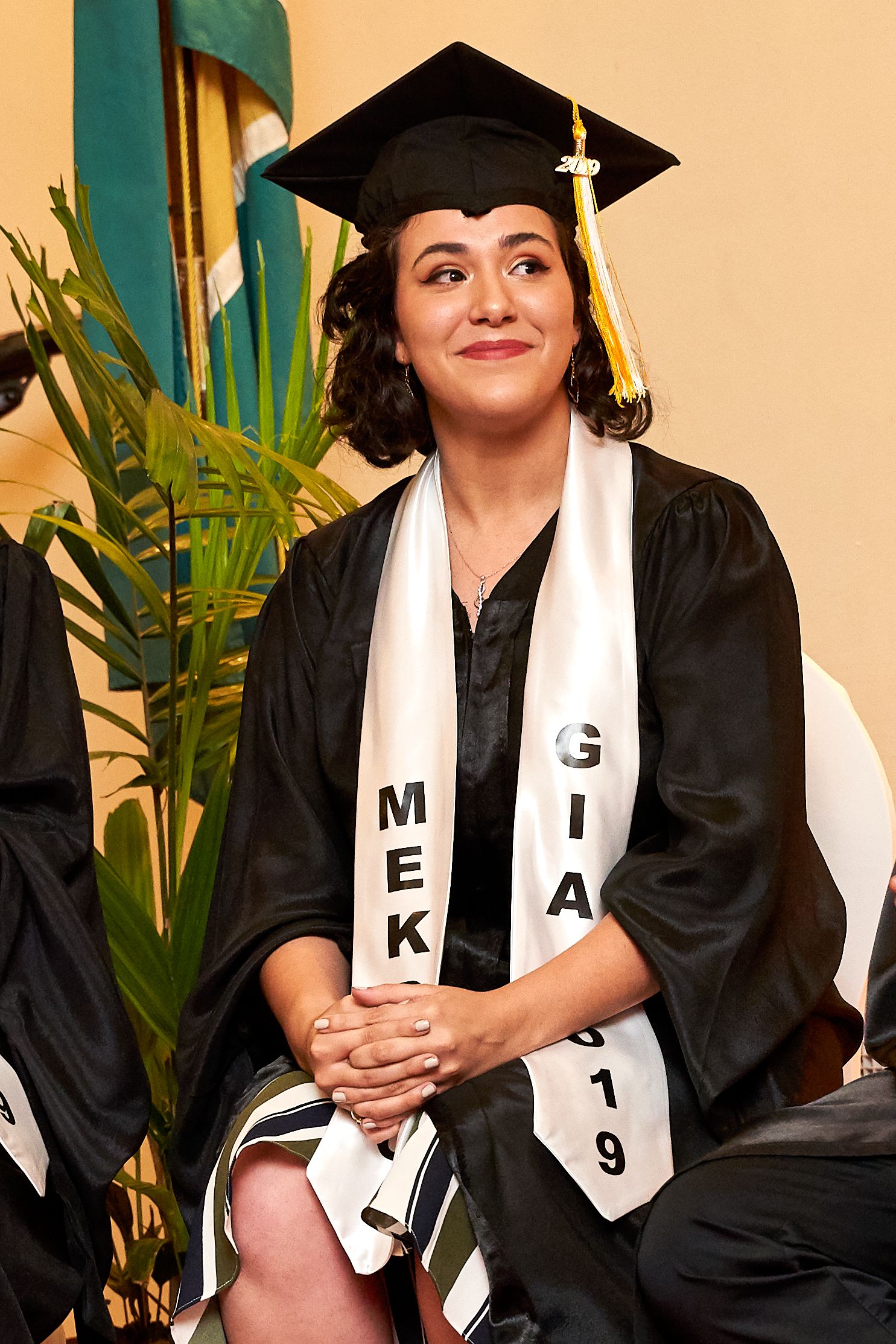 Graduate in black cap and gown with white sash smiles while posing in a series of photos against a tropical backdrop.